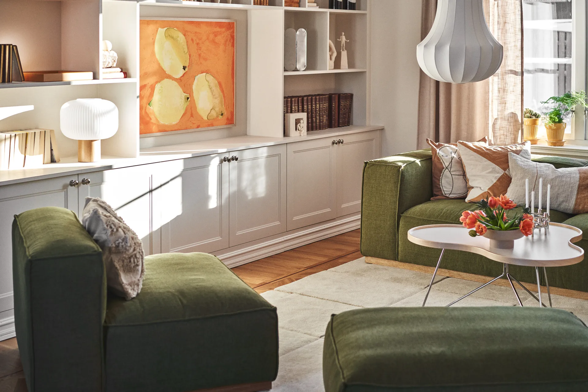 Salón soleado con sofás verdes, una mesa de centro blanca con tulipanes naranjas y velas, y estantes blancos con arte.