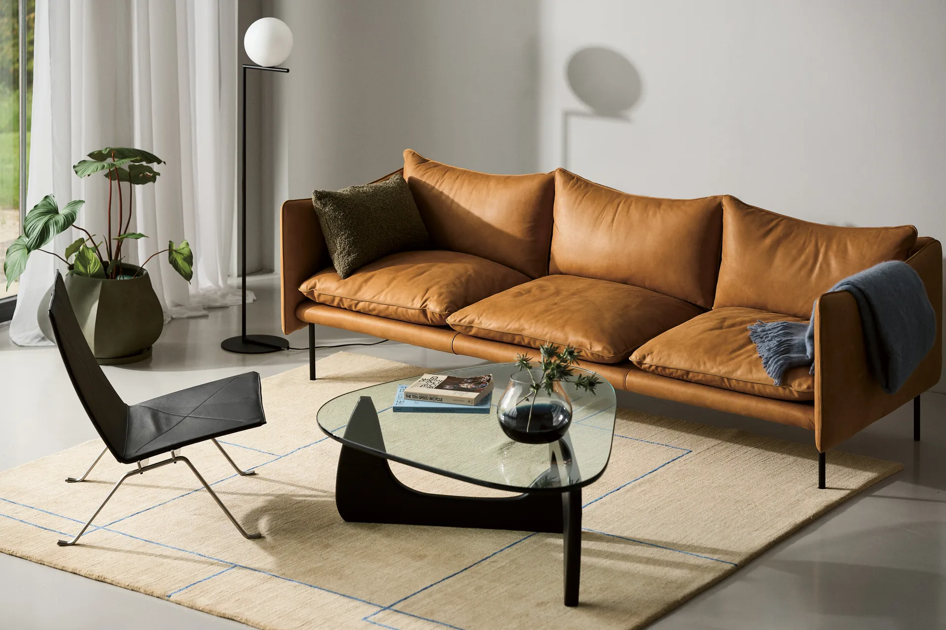 Una sala de estar moderna con sofá de cuero camel, sillón negro, mesa de centro de cristal y alfombra beige con líneas azules.