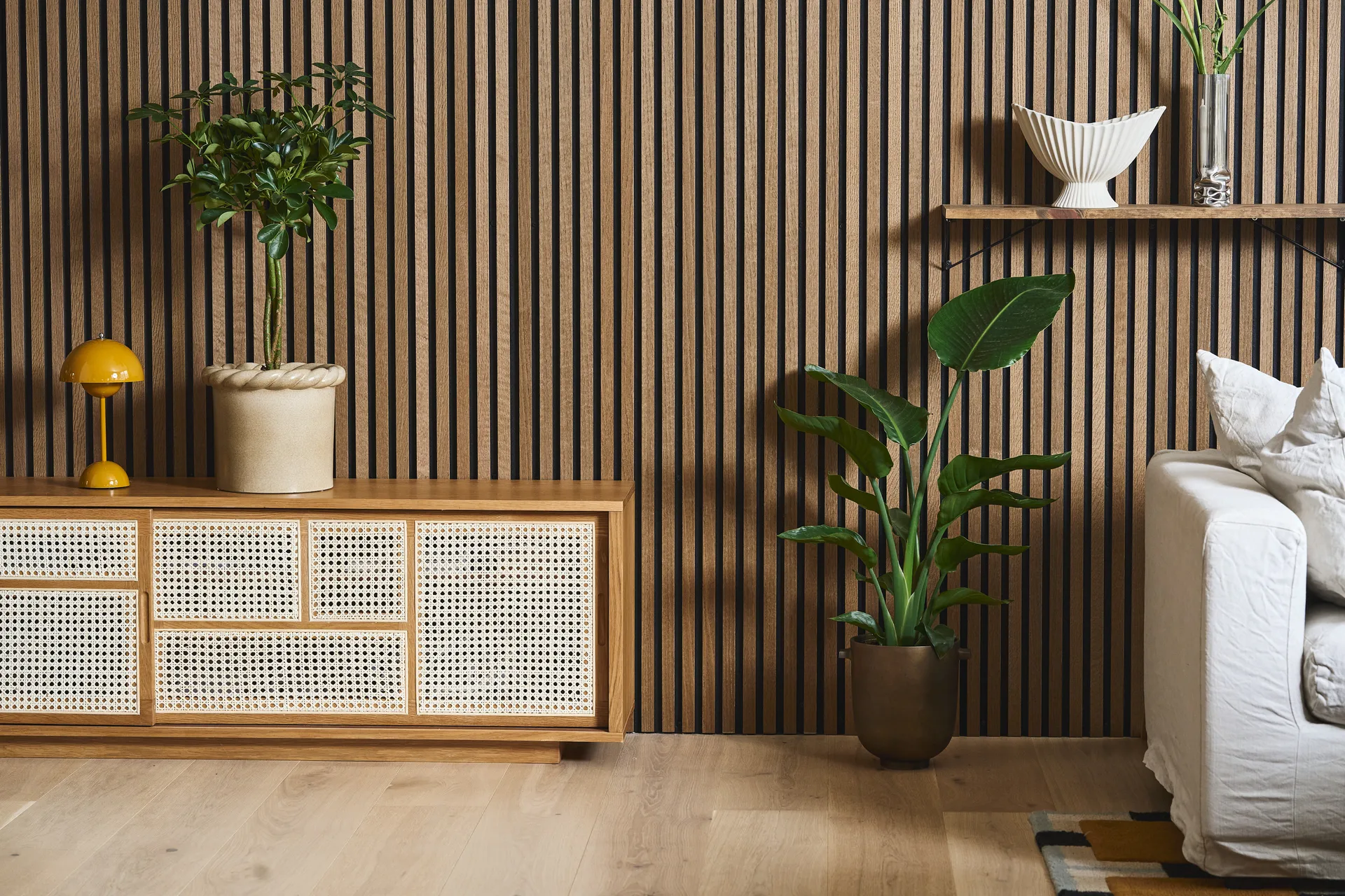 Salón moderno con pared de lamas de madera, aparador de ratán, lámpara amarilla, plantas y sofá blanco.