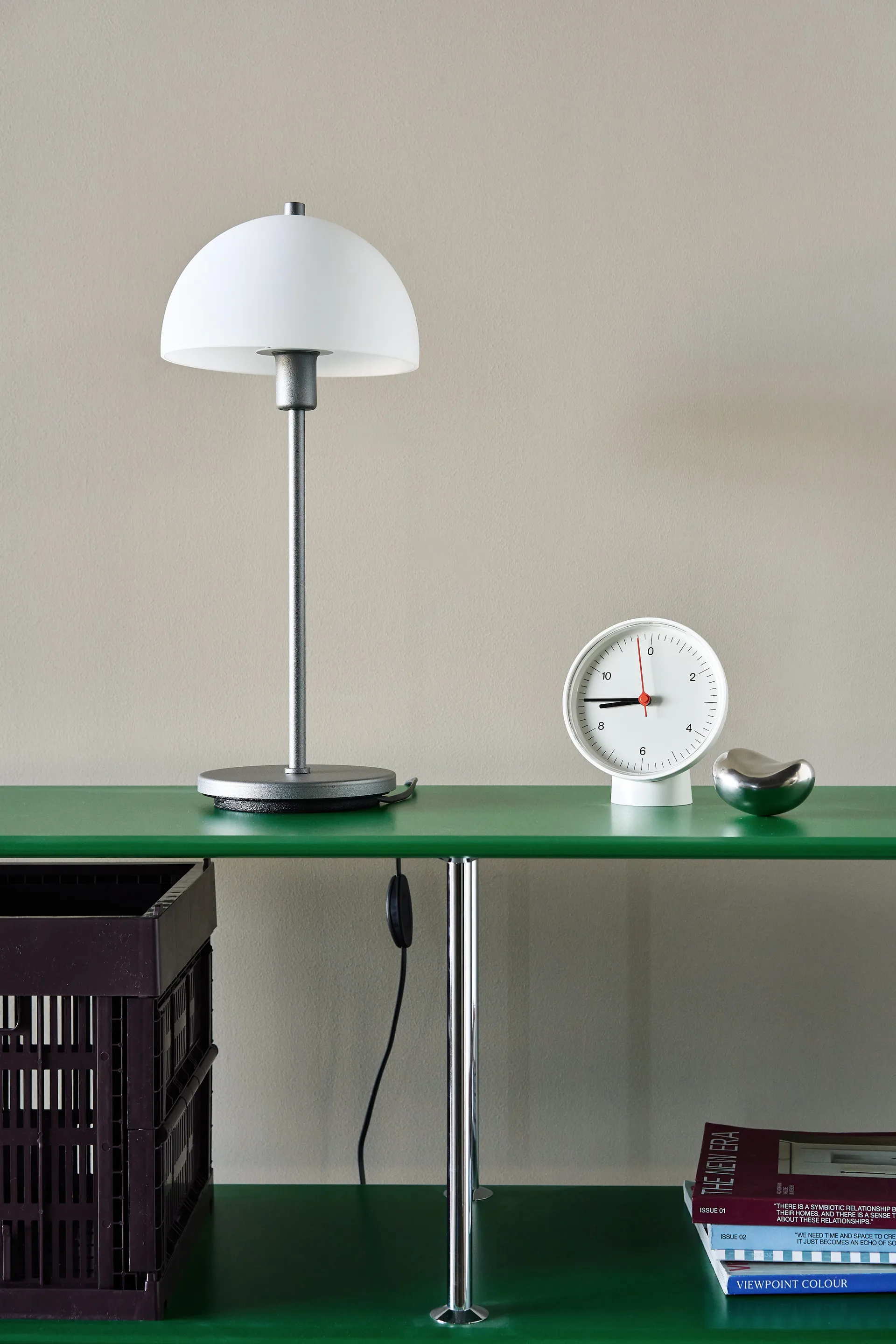 Estante verde minimalista con lámpara de cúpula blanca, reloj redondo blanco y guijarro plateado. Caja oscura y revistas debajo.