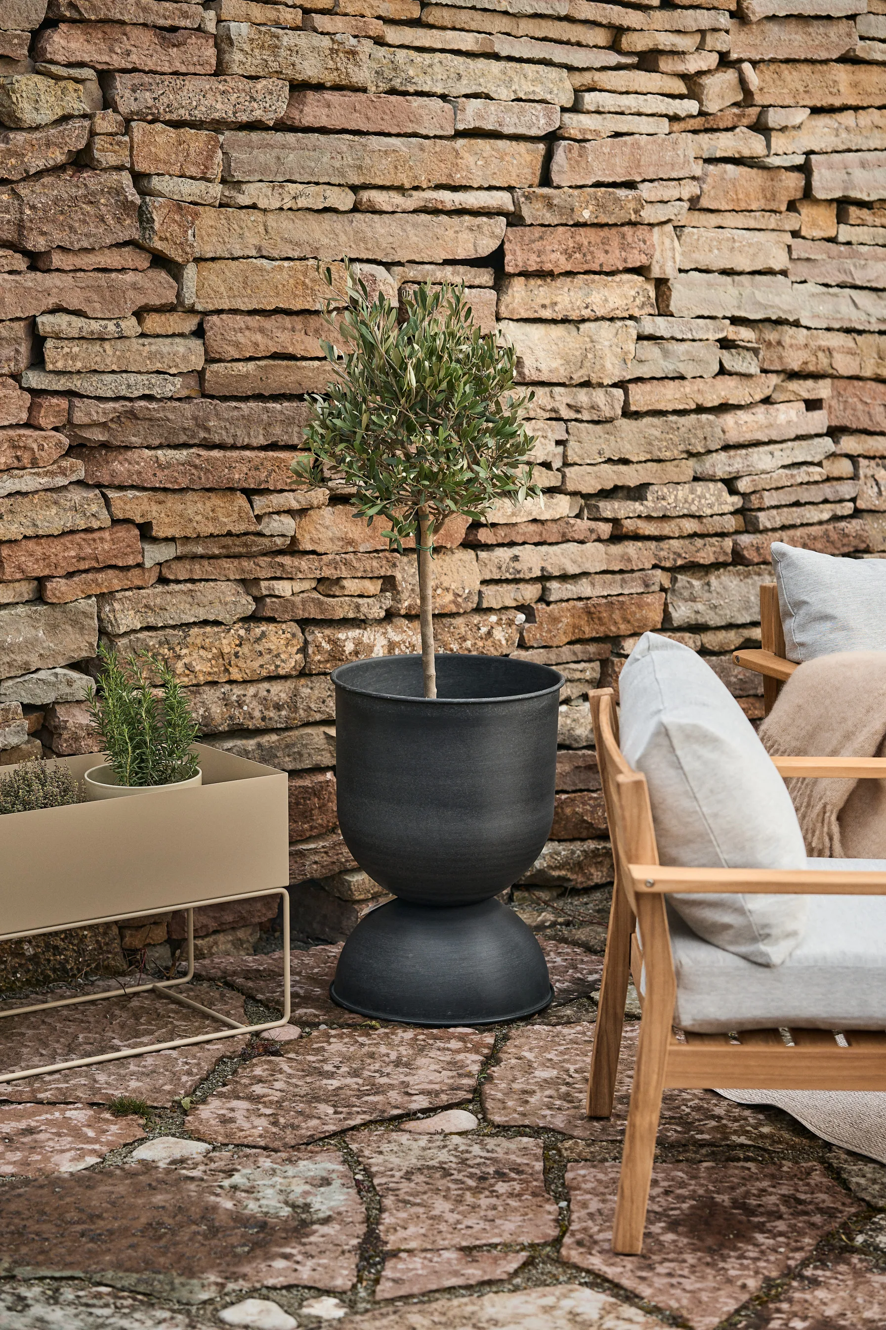 Pequeño árbol en una maceta negra con forma de reloj de arena y jardinera beige frente a un muro de piedra.