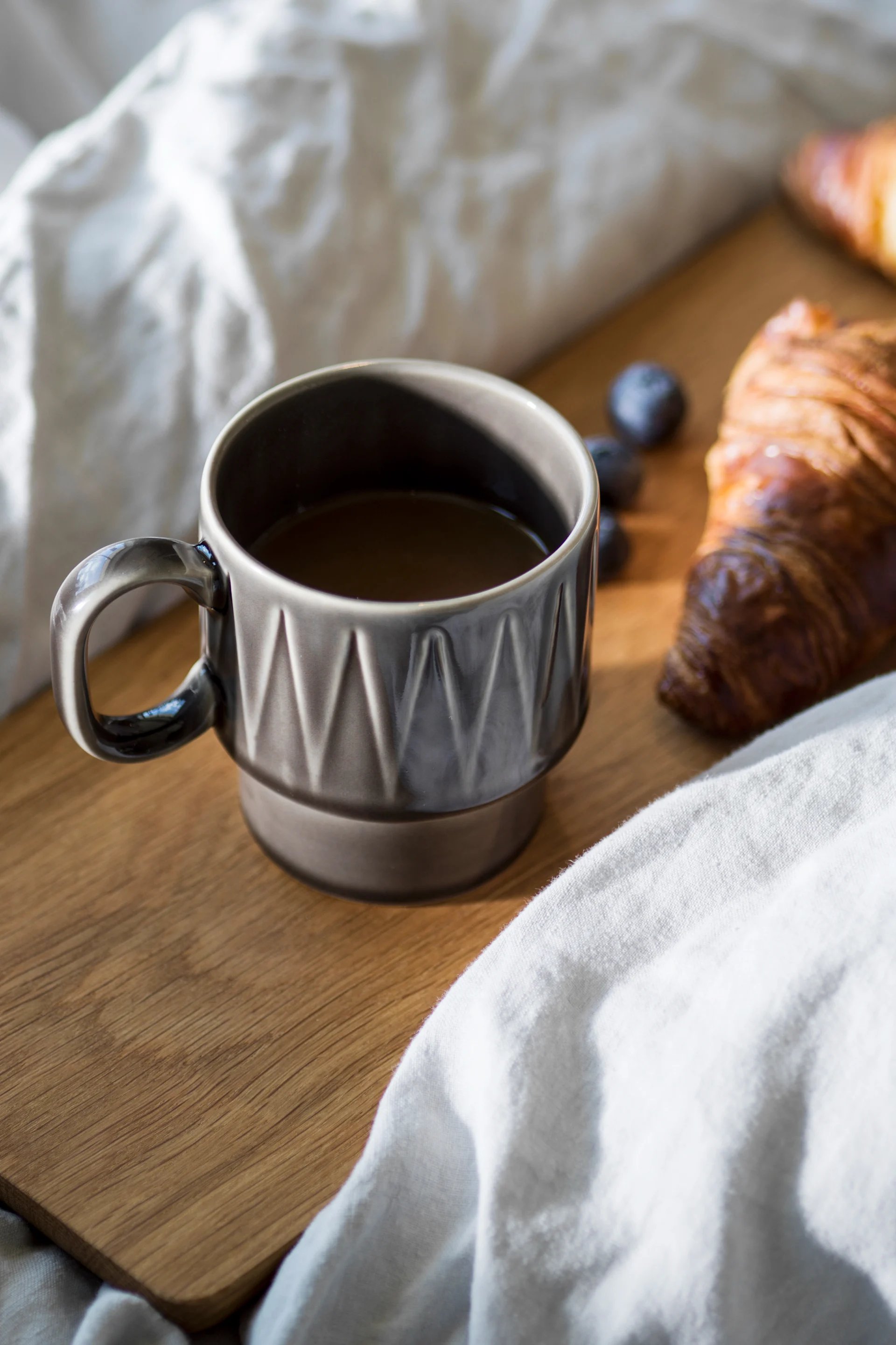 La taza de café espresso Coffee & More en gris está sobre una tabla de madera en una cama, con un croissant al lado.