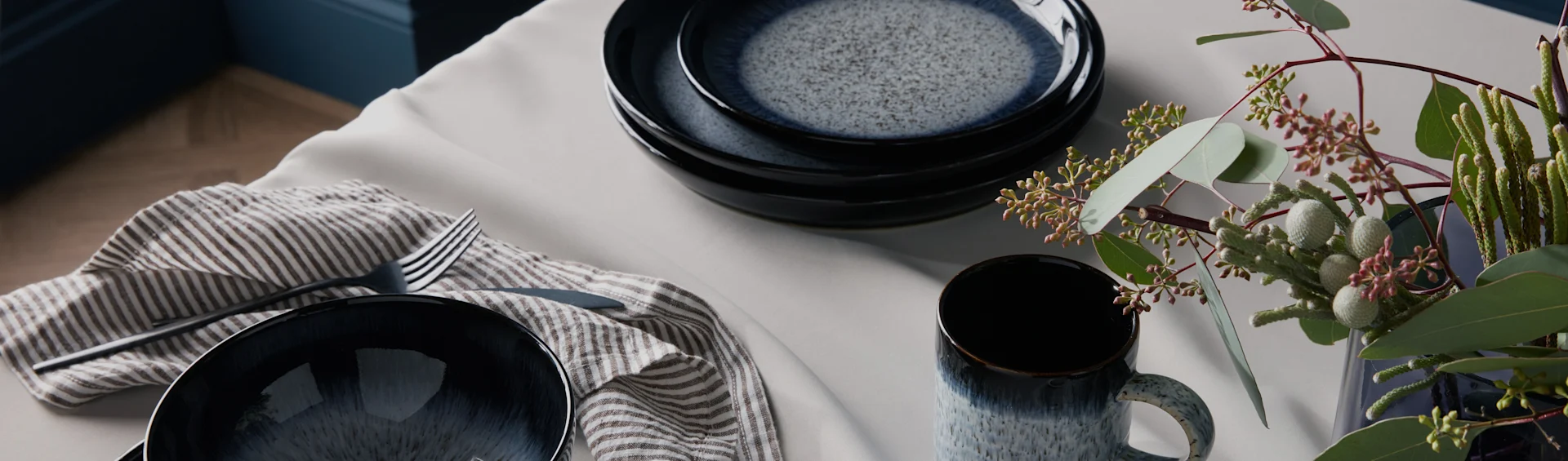 Vajilla de cerámica azul oscuro moteada, servilleta a rayas, cubiertos y follaje verde sobre una mesa clara.