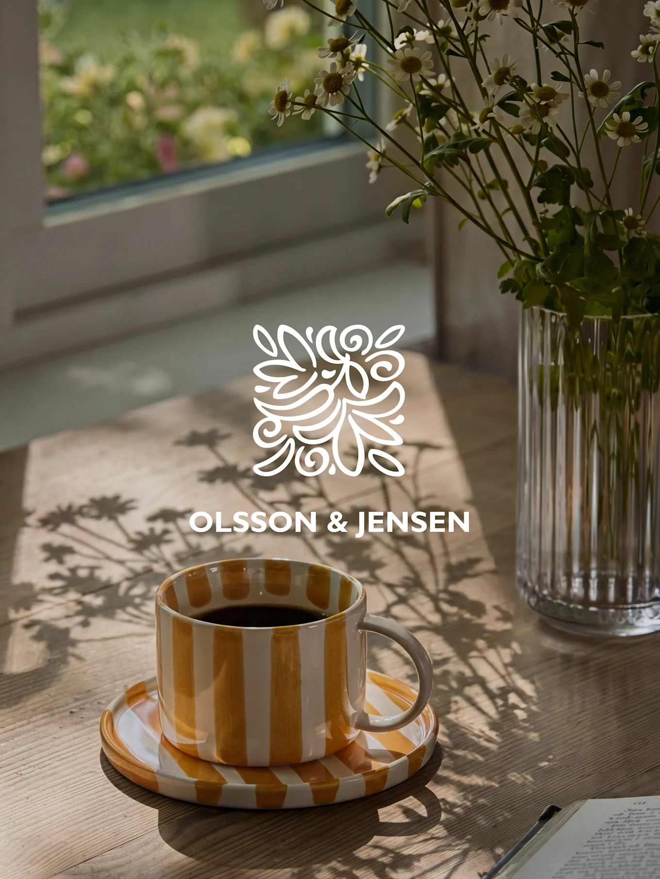 Taza de café a rayas amarillas y blancas en una mesa de madera con un jarrón de flores blancas y sombras solares, logo de Olsson & Jensen.