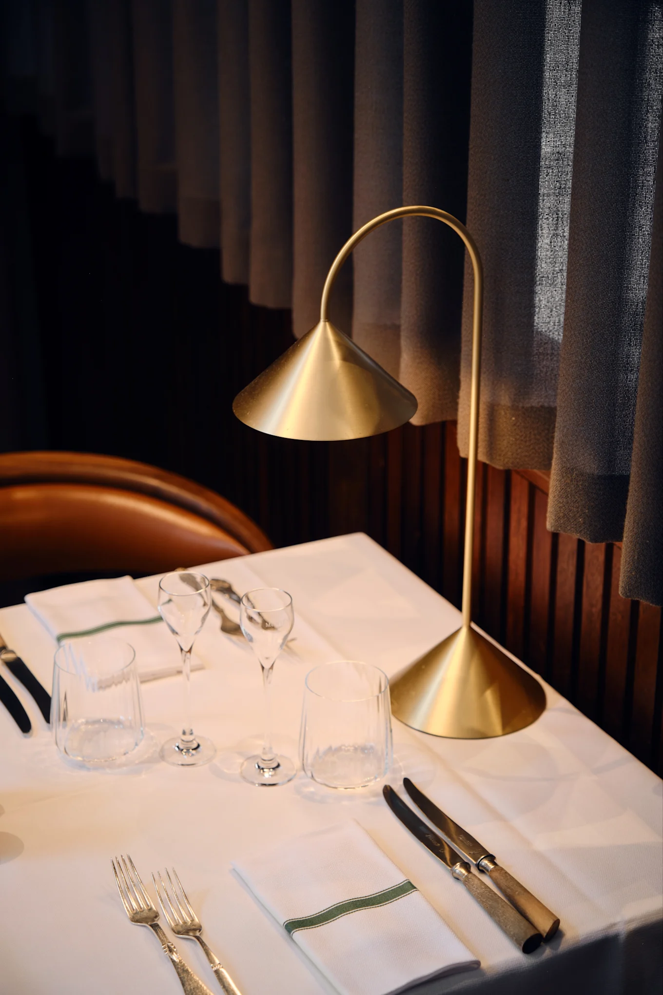 Una lámpara de mesa dorada moderna ilumina una mesa de restaurante puesta con mantelería blanca, vasos, cubiertos y una servilleta con una franja verde.