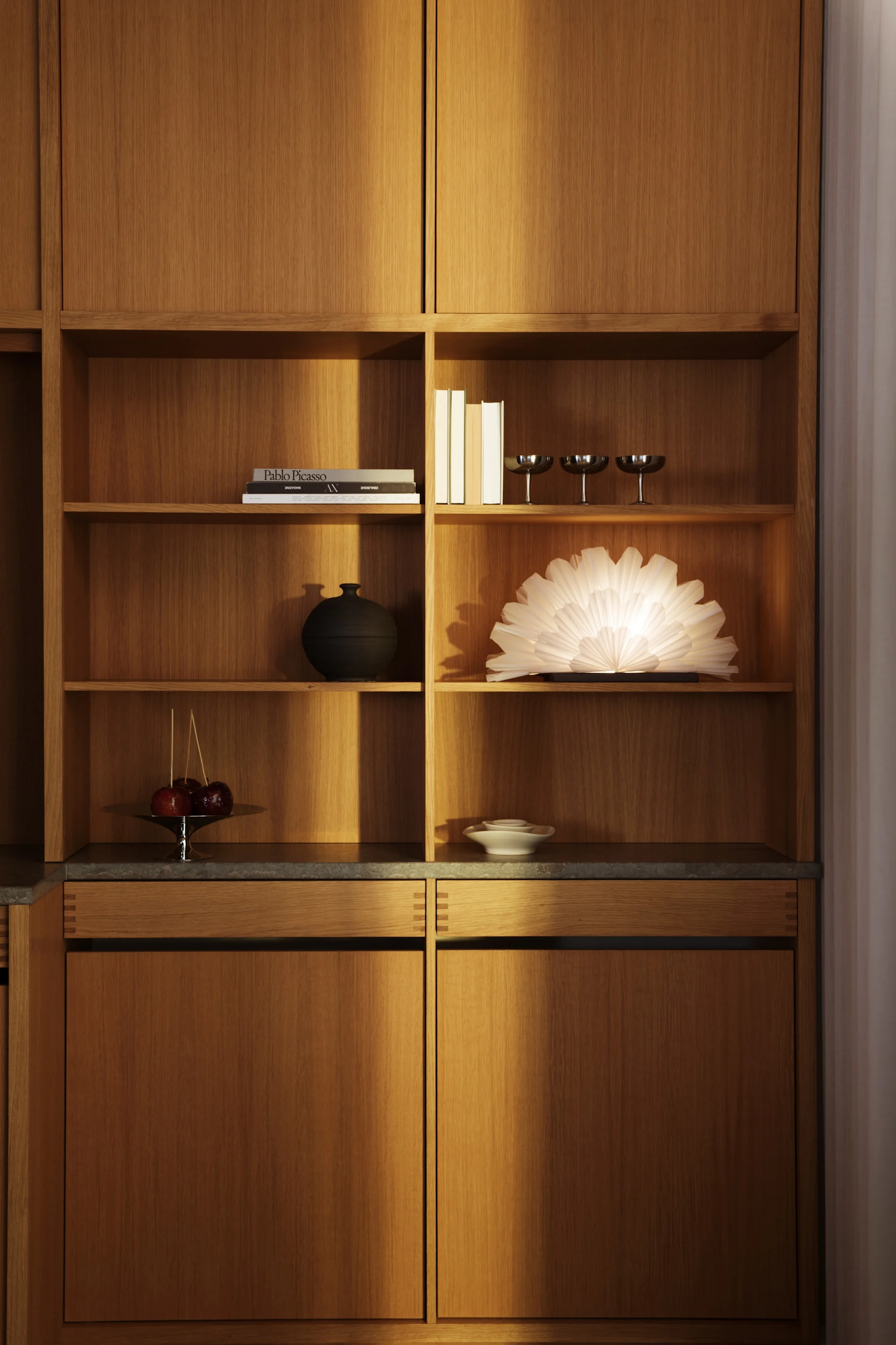 Una estantería de madera empotrada con diversos objetos decorativos, entre los que se incluyen libros, vasos de plata, un jarrón negro, una lámpara en forma de abanico de color blanco brillante, manzanas caramelizadas y cuencos blancos.