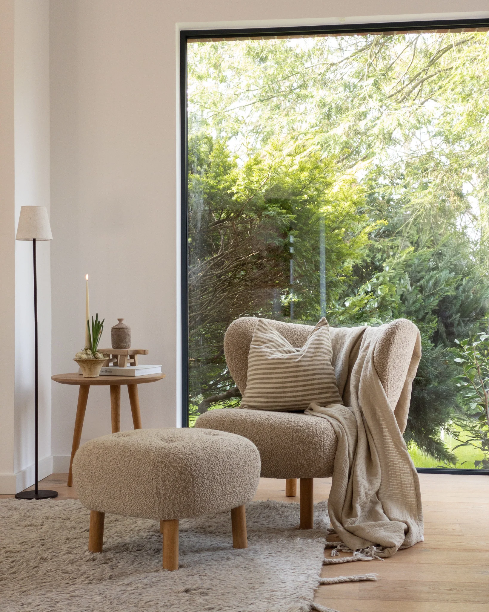Rincón acogedor: sillón marrón claro, otomana, mesa auxiliar con vela, ventana grande, árboles verdes.