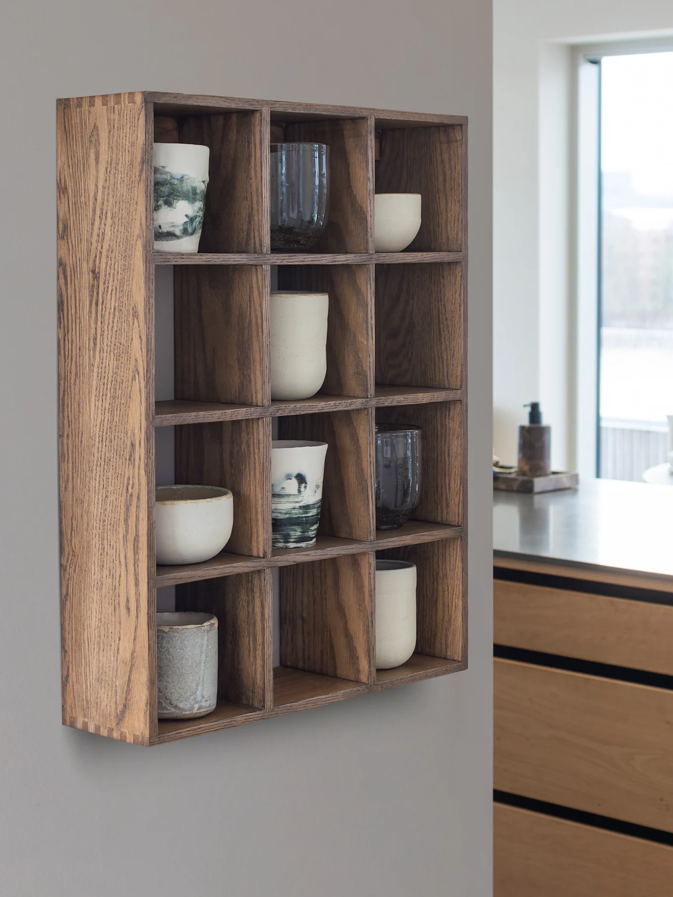 Estante de pared de madera con 12 compartimentos que contienen tazas y vasos de cerámica. Una cocina moderna y una ventana están en el fondo.