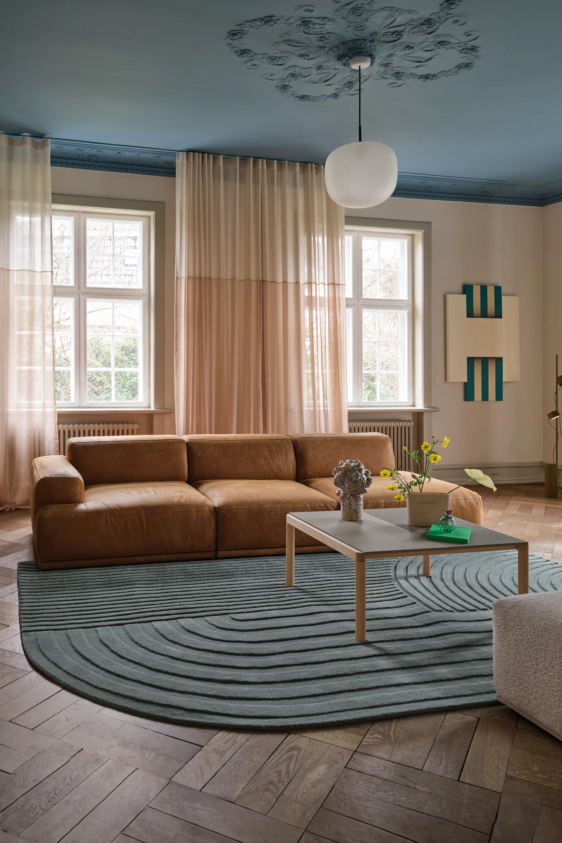 Sala de estar moderna: sofá de cuero marrón, alfombra turquesa con patrón, techo azul claro con lámpara colgante, ventanales.