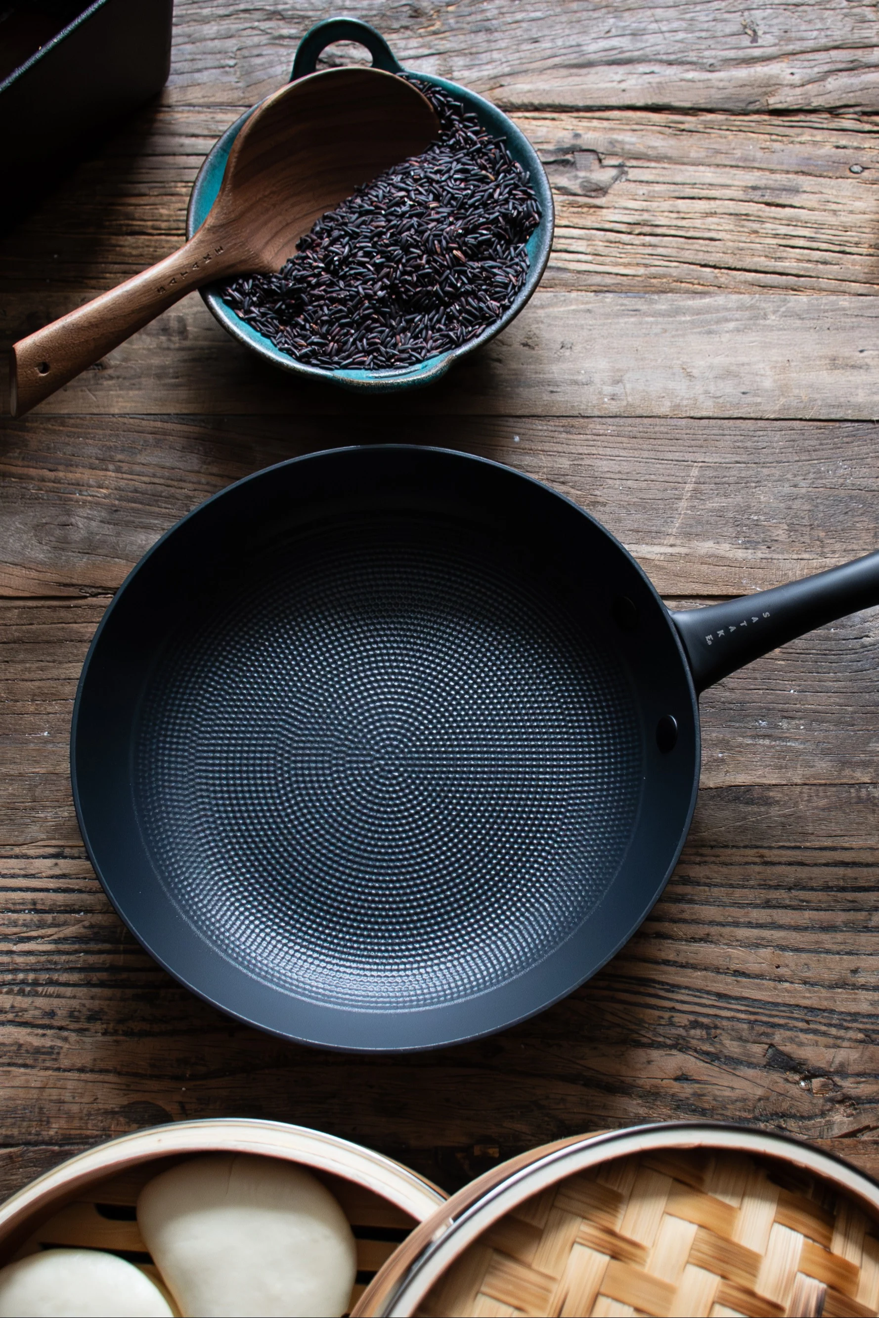 Sartén negra, cuenco con arroz negro y cuchara de madera, vaporeras de bambú con moños blancos sobre una mesa de madera rústica.