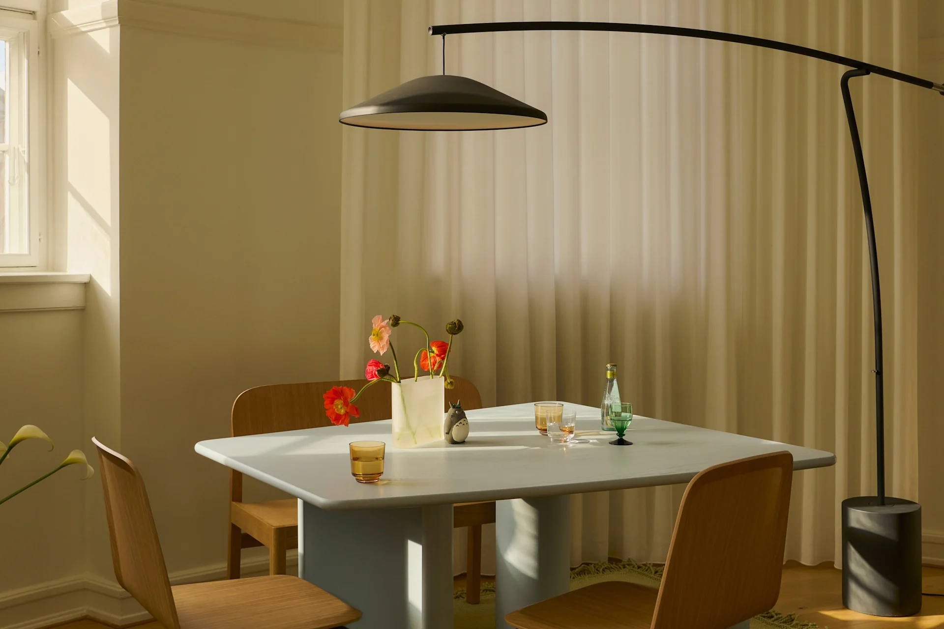 Comedor moderno con mesa azul claro, sillas de madera, lámpara de pie negra y jarrón de flores coloridas al sol.