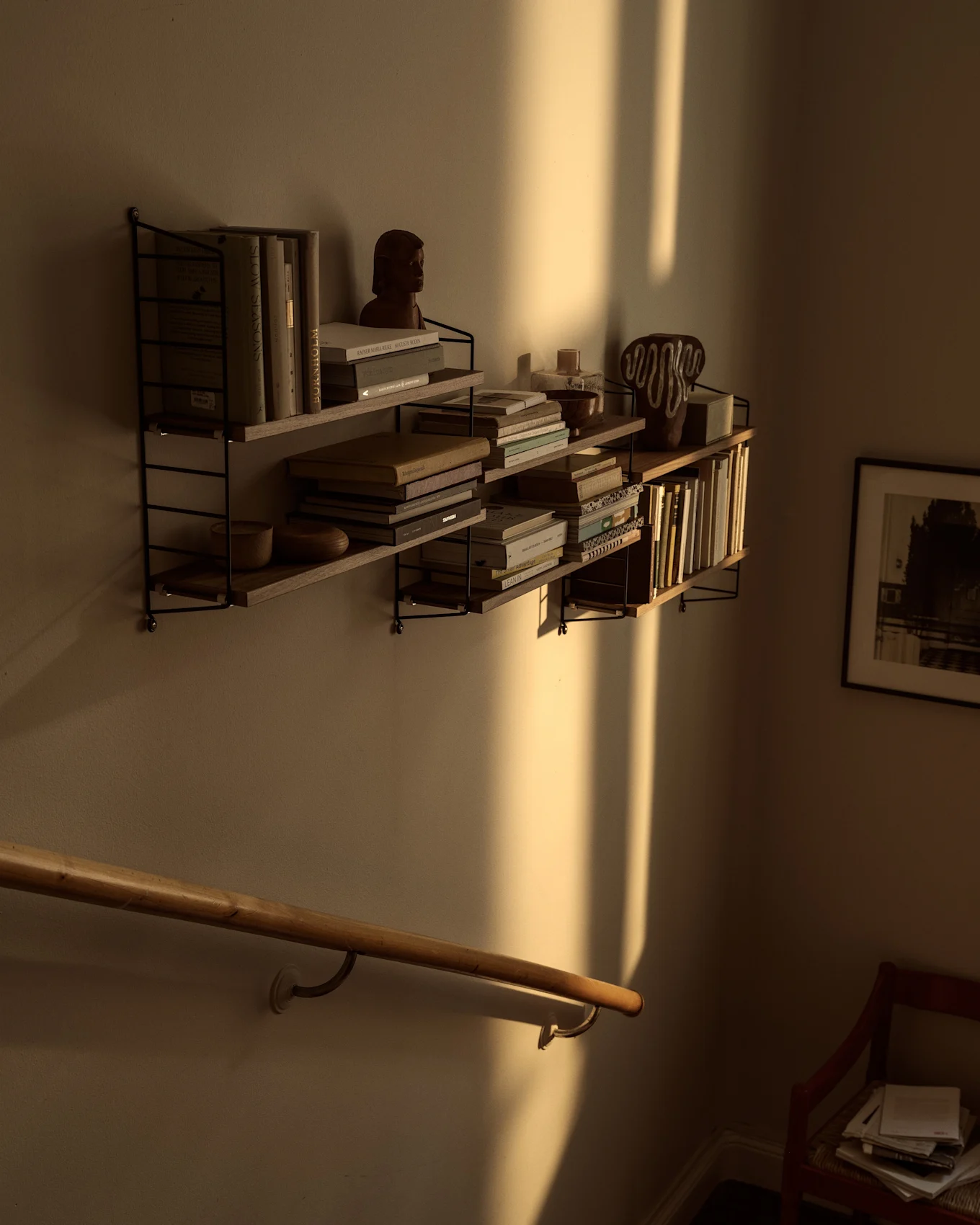 Estantes de pared con libros y decoración, bañados por el sol cálido que proyecta sombras verticales sobre un pasamanos de madera.