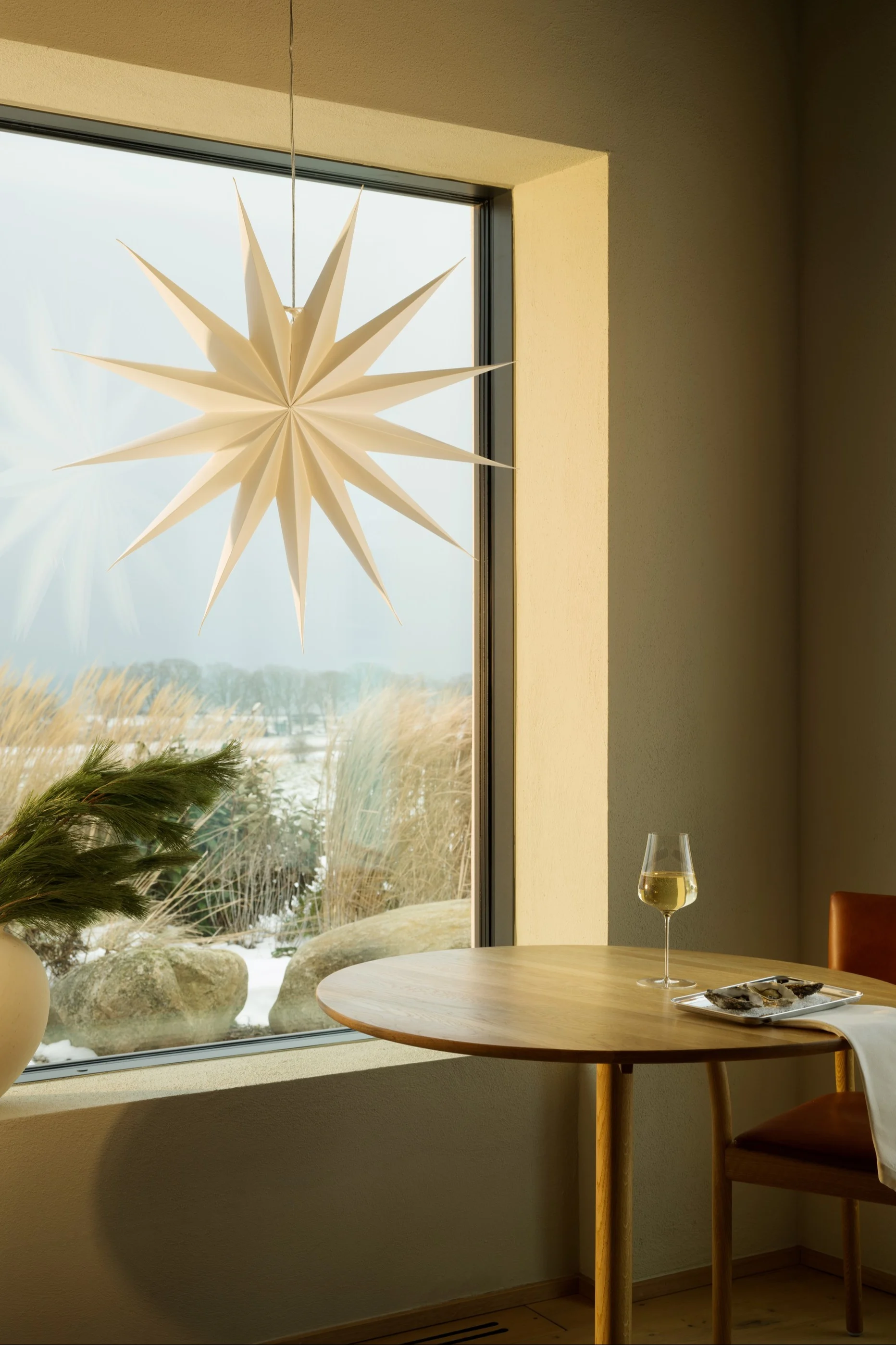 Una ventana da a un paisaje nevado en el que se ve una linterna en forma de estrella. Sobre una mesa hay vino blanco, ostras y una silla de cuero marrón.