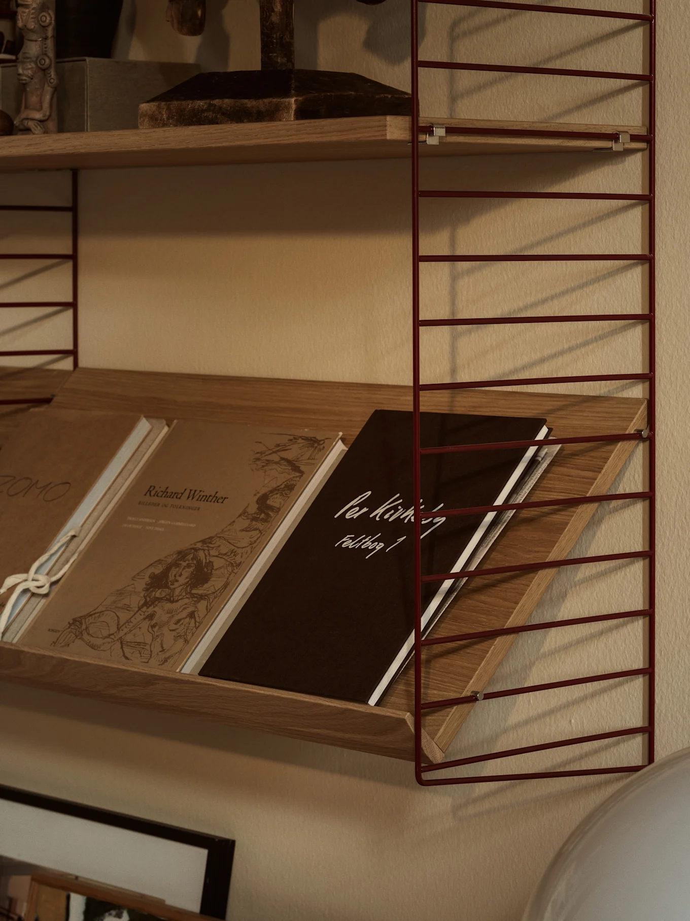Una estantería de alambre de metal rojo con estantes de madera. El estante inferior tiene libros inclinados, incluido uno marrón oscuro con 'Per Kirkeby Feldbog 1'.