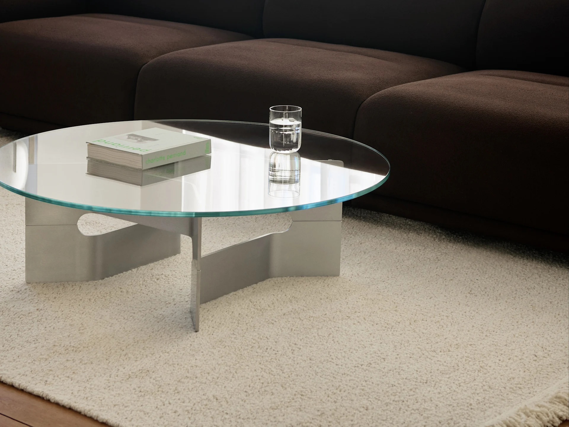 Mesa de centro redonda de cristal con libros y un vaso de agua sobre una alfombra de pelo largo color crema frente a un sofá marrón oscuro.