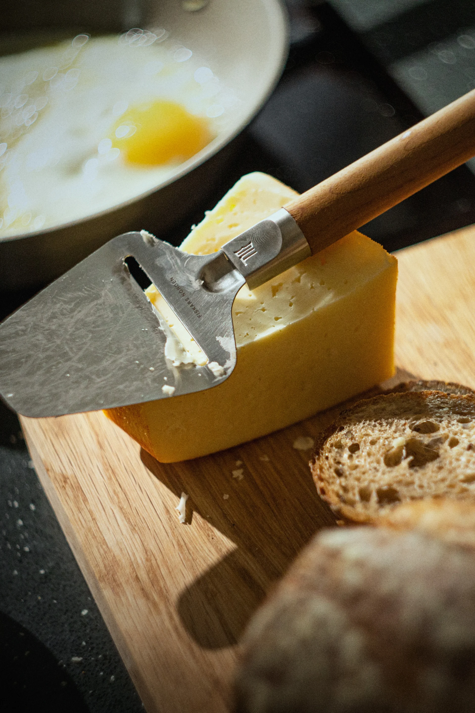 Un cortador de queso reposa sobre un bloque de queso amarillo en una tabla de madera, con rebanadas de pan y un huevo frito en una sartén al fondo.