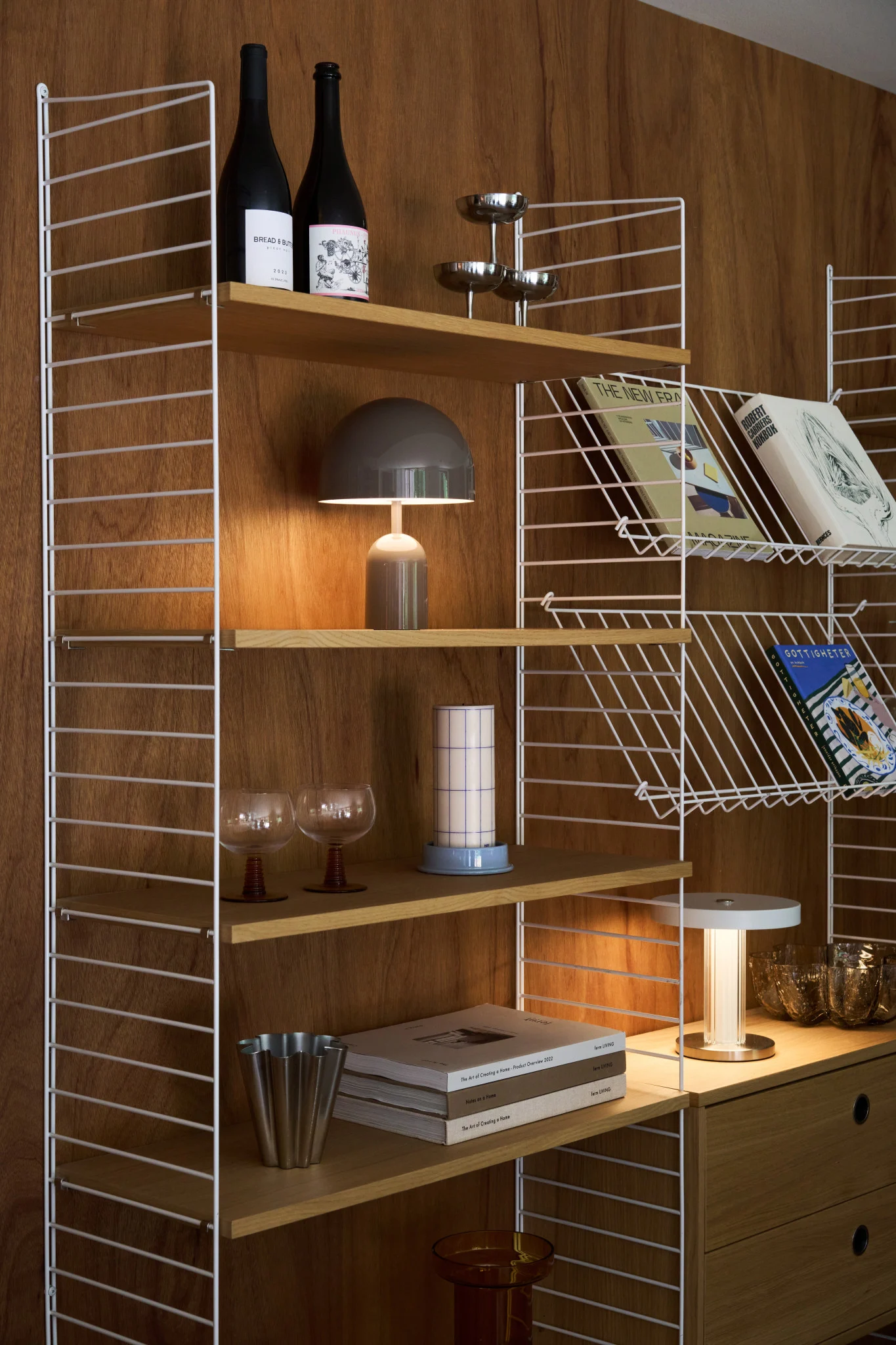 Una elegante estantería de madera con marcos de alambre blancos contiene libros, botellas, una lámpara de seta y varios artículos decorativos.