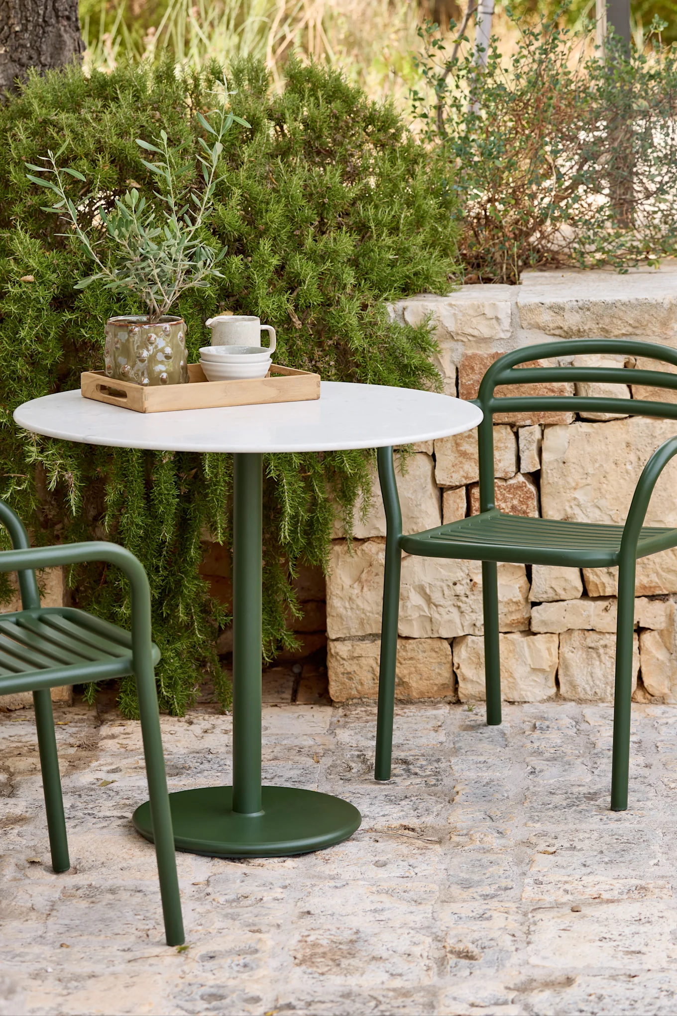 Un conjunto de comedor exterior con una mesa redonda blanca y dos sillas de metal verde oscuro en un patio de piedra, rodeado de exuberante follaje verde.