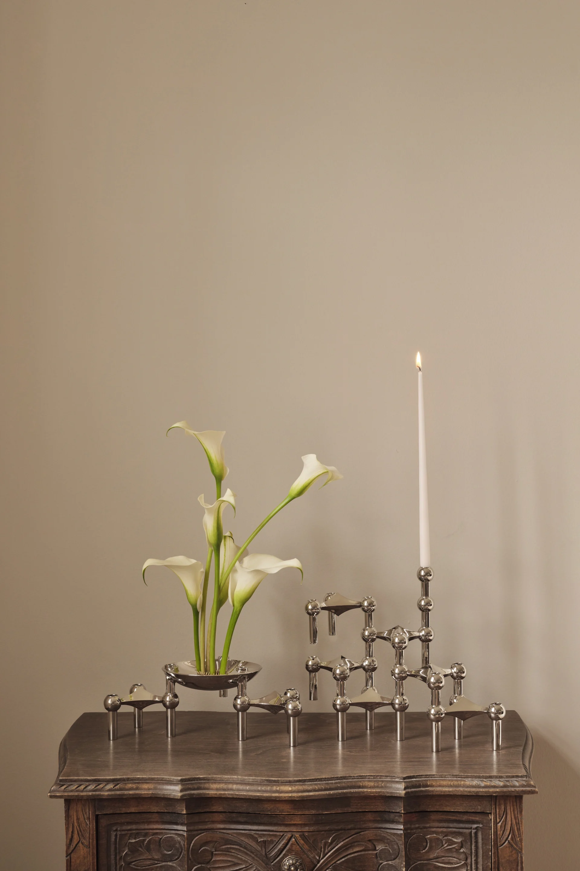 Lirios cala blancos en un cuenco plateado junto a un candelabro modular plateado con una vela blanca encendida sobre una mesa de madera tallada.