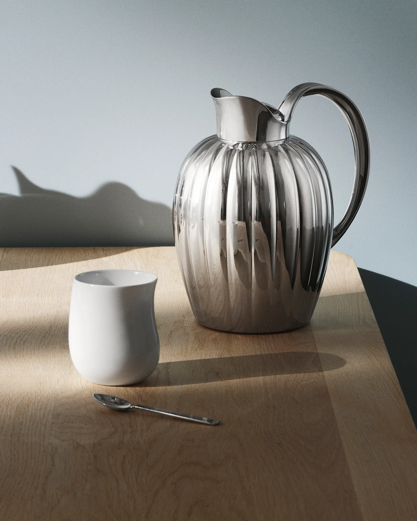 Dos jarras de plata acanaladas, una taza blanca y una cuchara de plata sobre una mesa de madera contra una pared azul-gris.