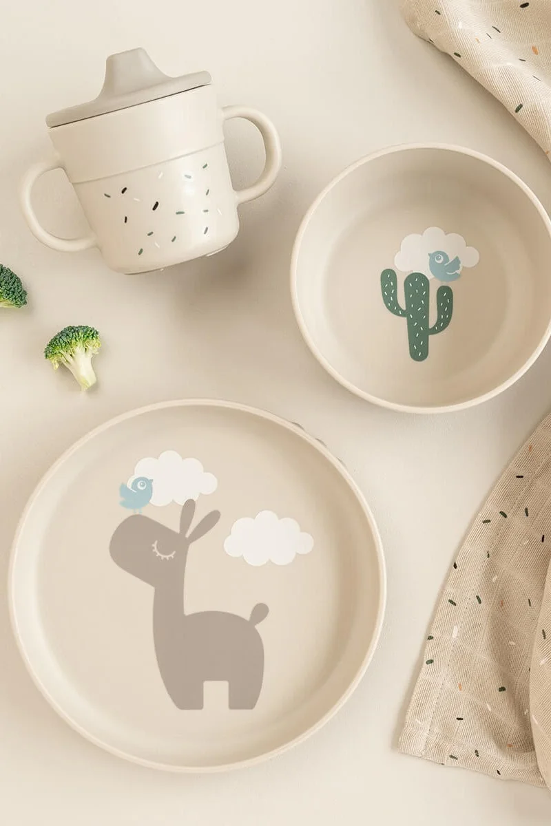 Un juego de vajilla infantil beige claro con una taza con boquilla, un cuenco con un cactus y un plato con una llama y pájaros.