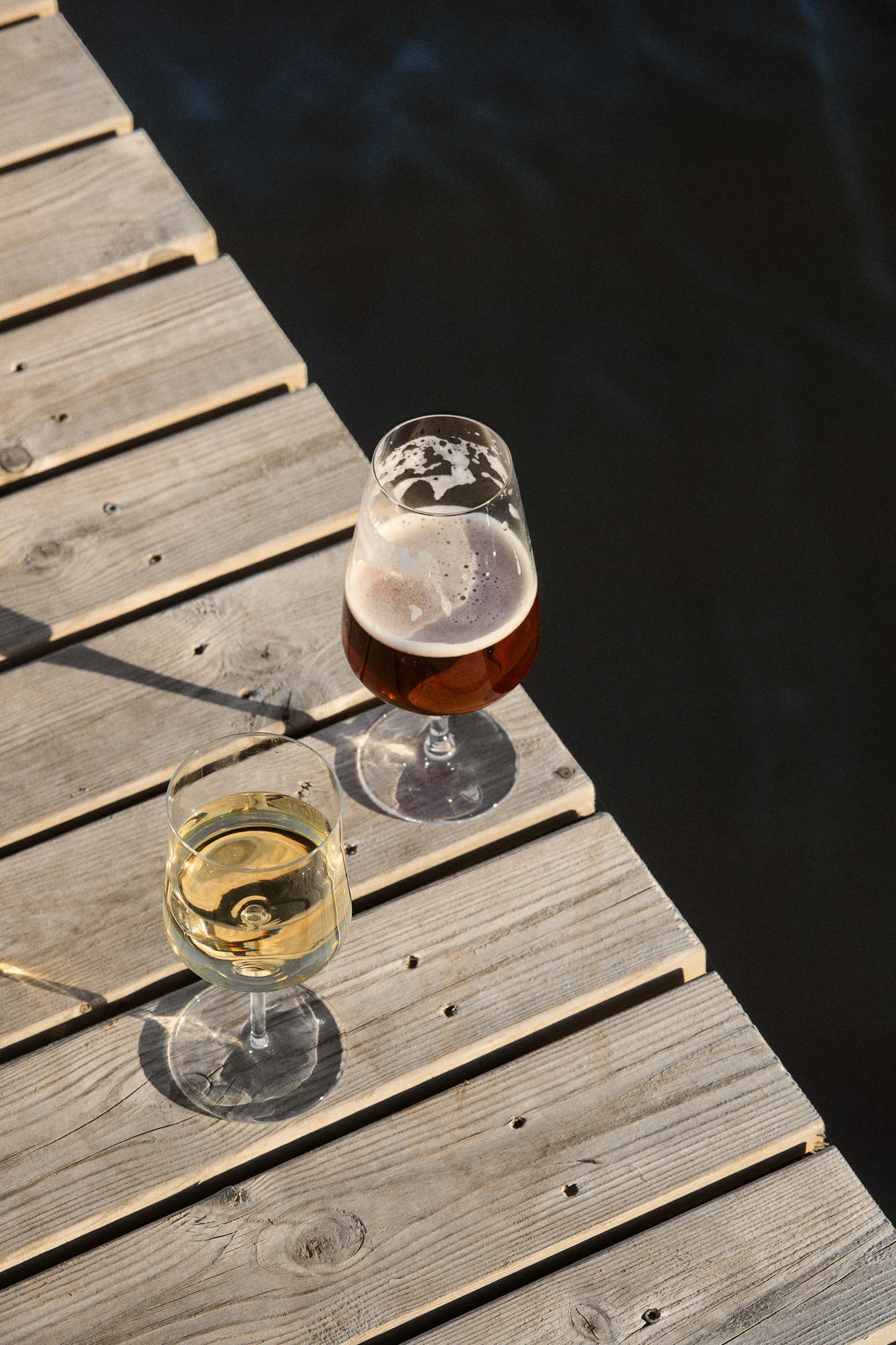 Dos copas, una de vino blanco y otra de cerveza, en un muelle de madera junto a agua oscura bajo el sol.