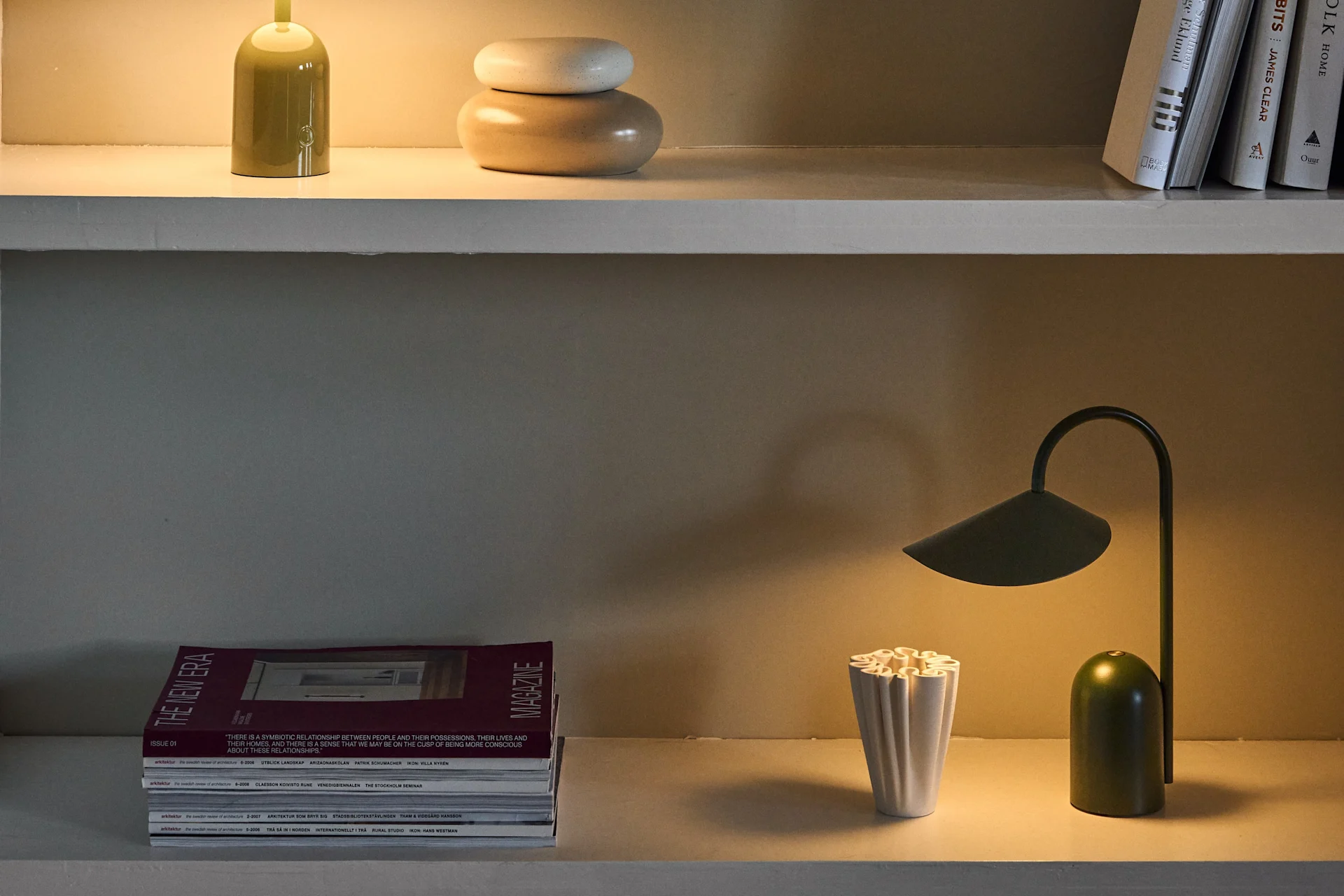 Tres estantes muestran varias lámparas de mesa en verde y morado, junto con libros, revistas y objetos decorativos.