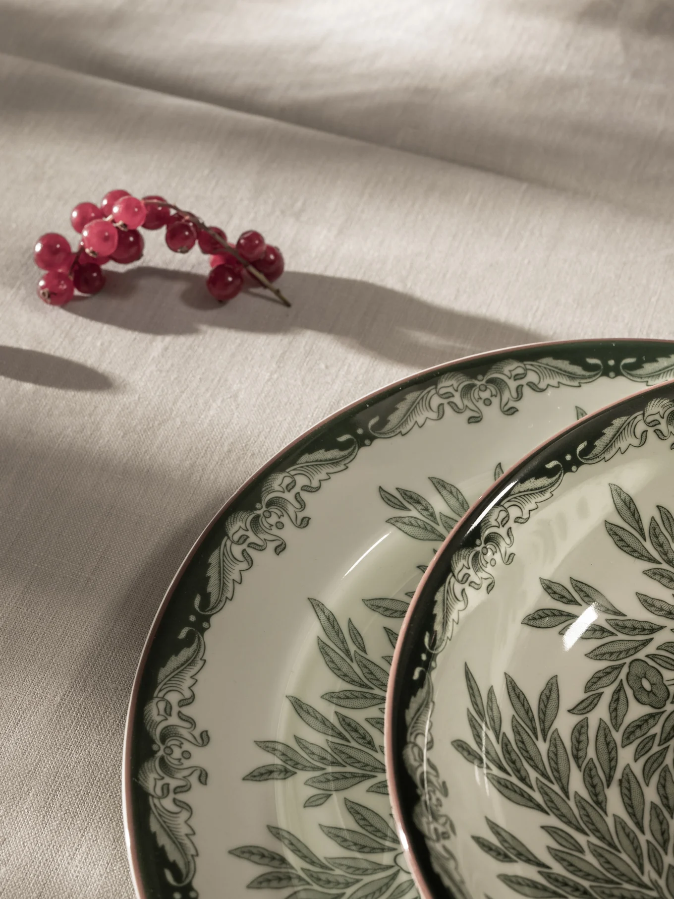 Grosellas rojas sobre un mantel claro junto a dos platos blancos con motivos de hojas verdes y bordes rosados.