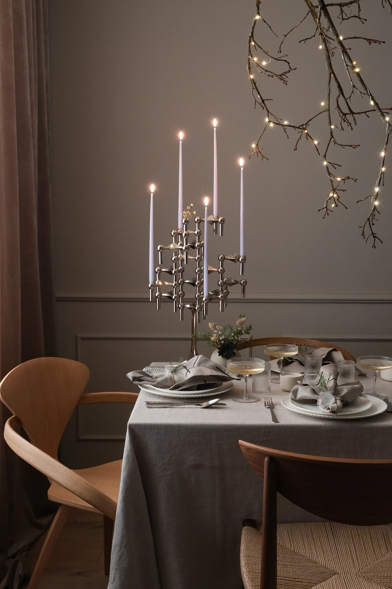 Una mesa de comedor puesta para una ocasión especial con un candelabro moderno, vajilla y ramas iluminadas.