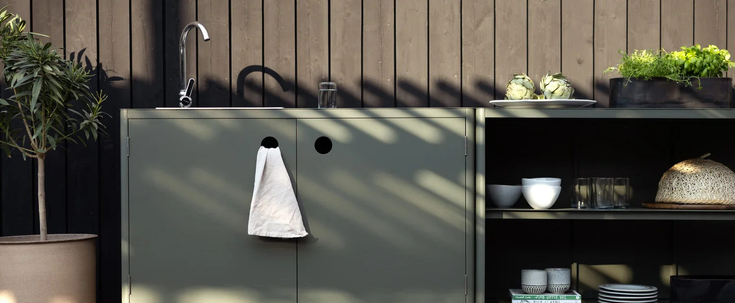 Cocina exterior con gabinetes verde salvia, fregadero, hierbas y una planta en maceta contra una pared de madera oscura.