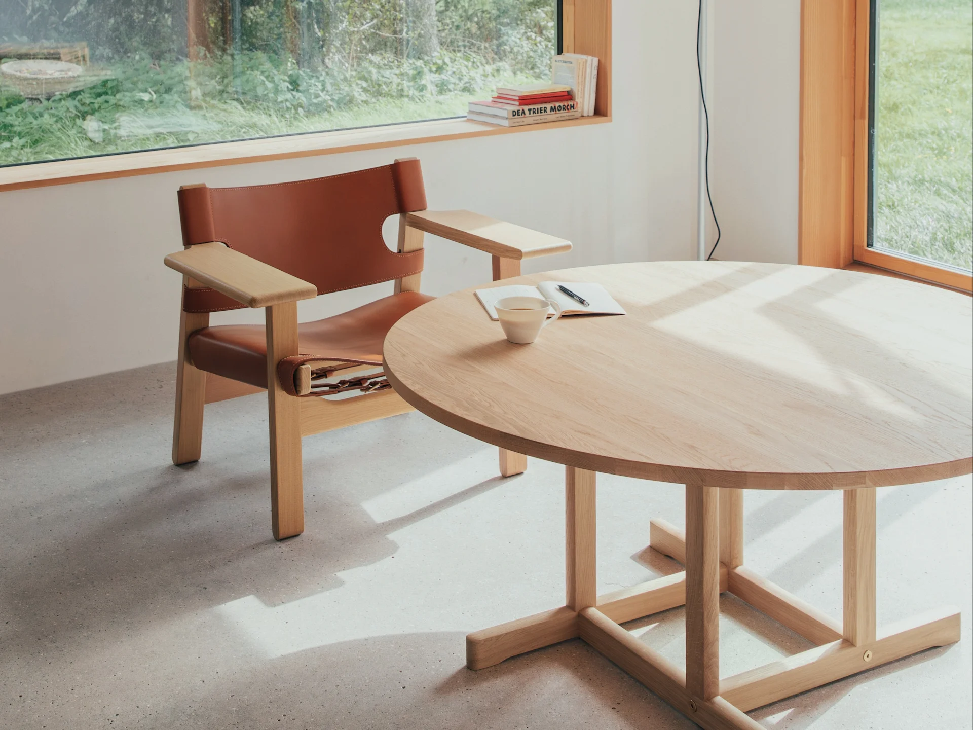 Habitación luminosa con sillón de cuero marrón, mesa redonda de madera clara con taza y cuaderno, iluminada por ventana con vistas a la vegetación.