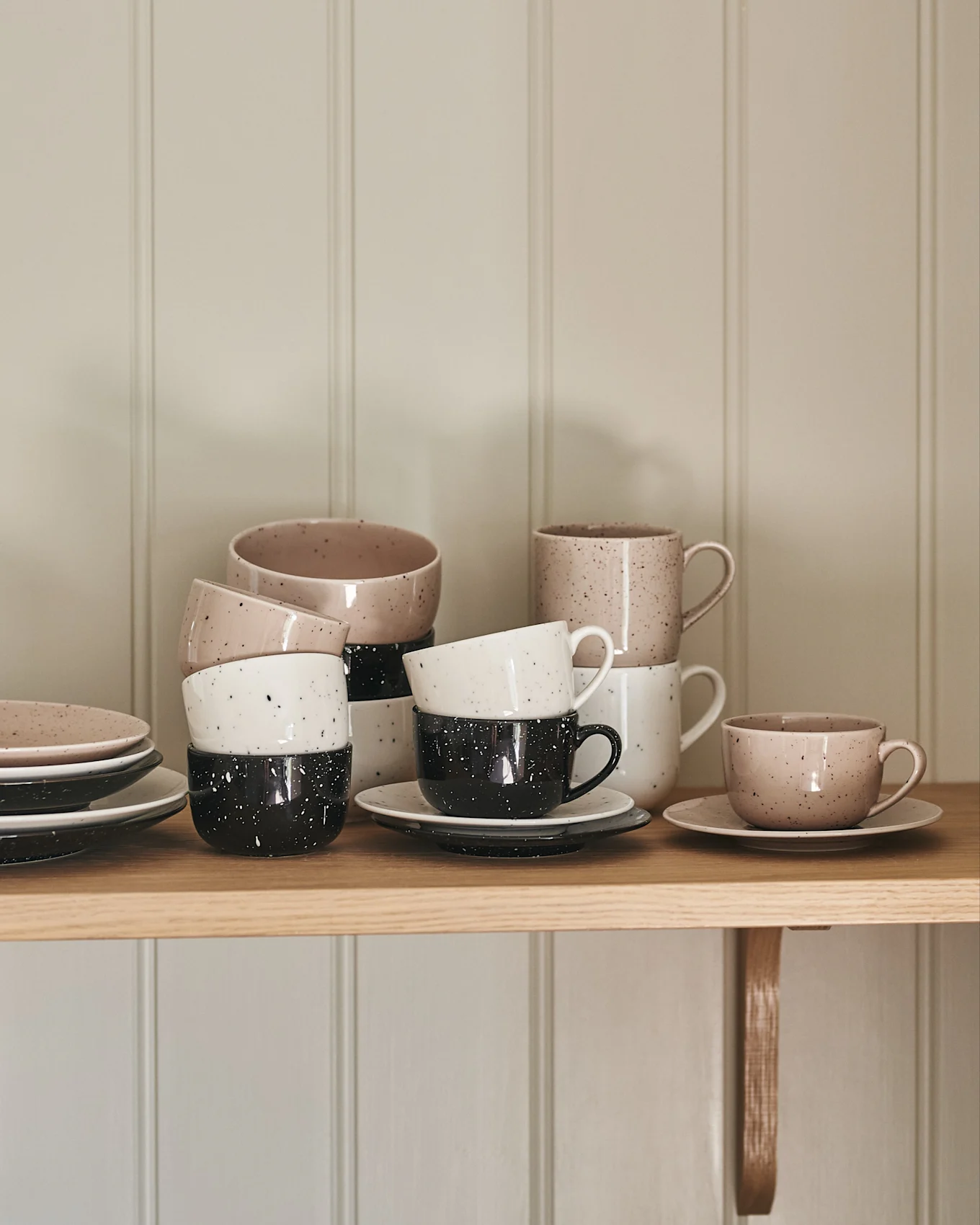 Tazas y platos de cerámica apilados en beige, blanco y negro con salpicaduras sobre un estante de madera.