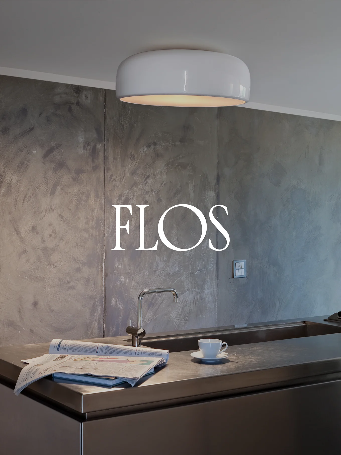 Cocina moderna con luz de techo blanca y redonda, encimera de acero inoxidable con fregadero, periódico y taza de té. Logotipo de FLOS en pared texturizada.