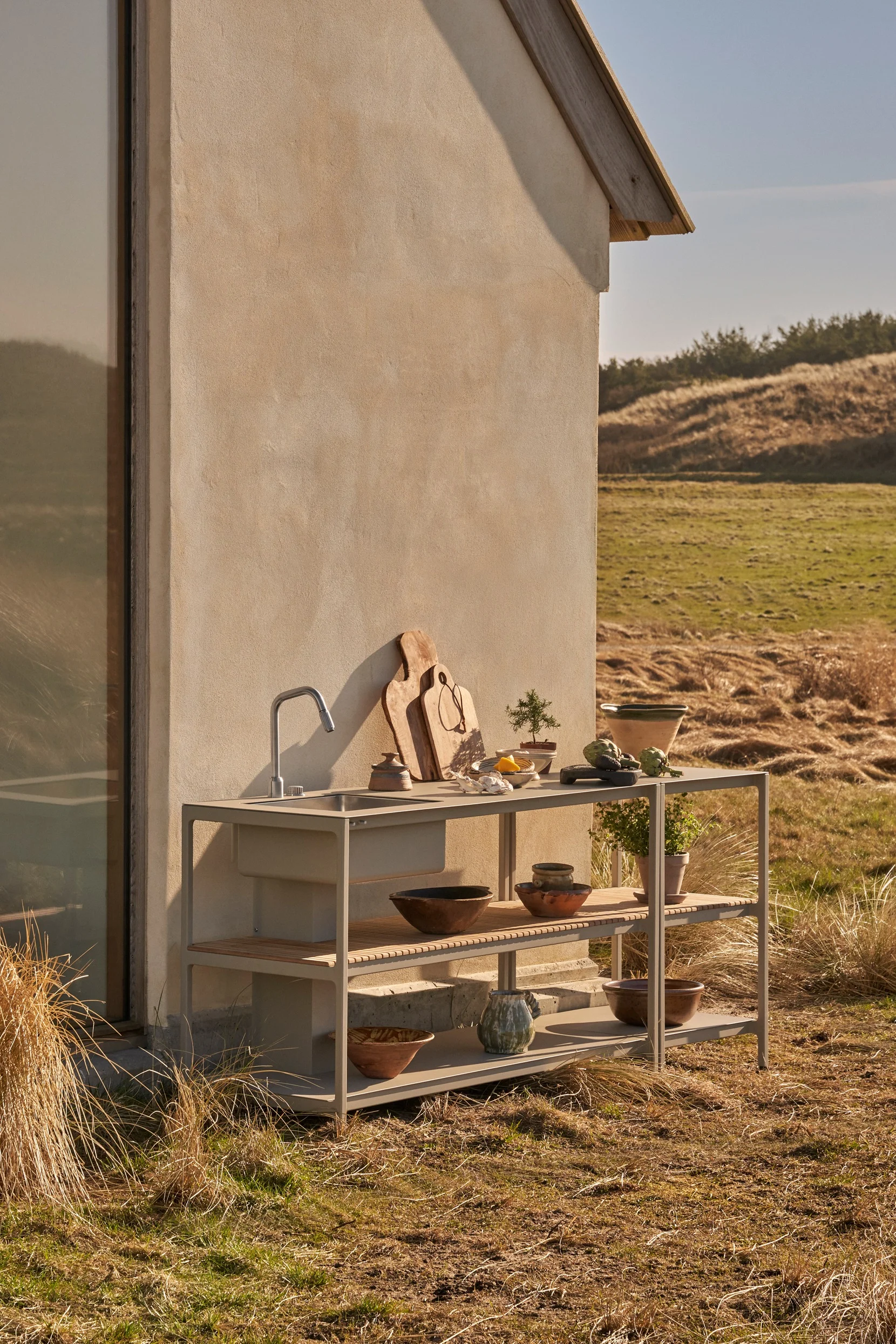 Módulo de cocina exterior con fregadero y estantes, decorado con tablas de cortar, plantas y cuencos, frente a un edificio en un paisaje natural.