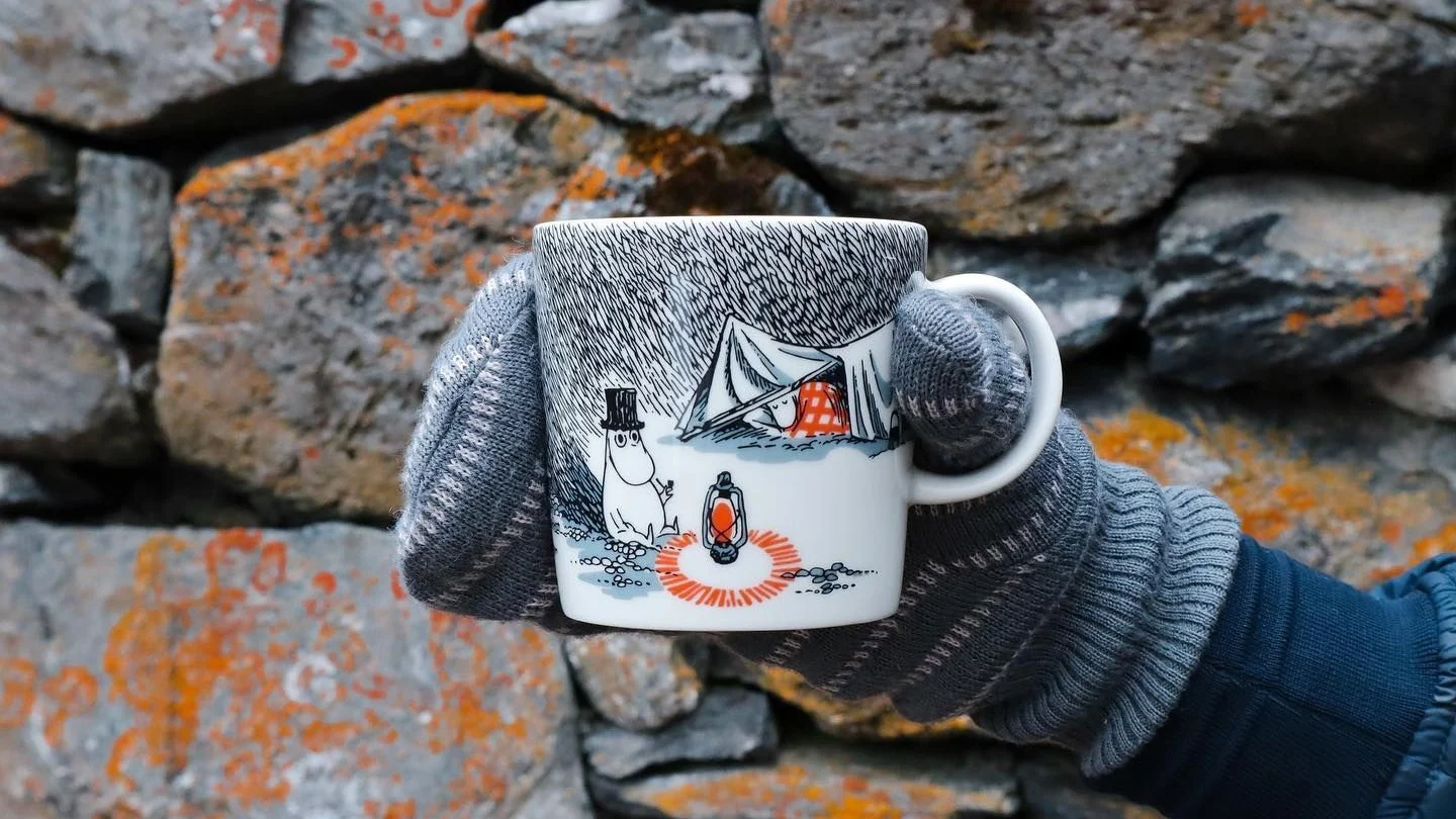 @moominbyingri sostiene la taza de Mumin delante de un muro de piedra con un guante.