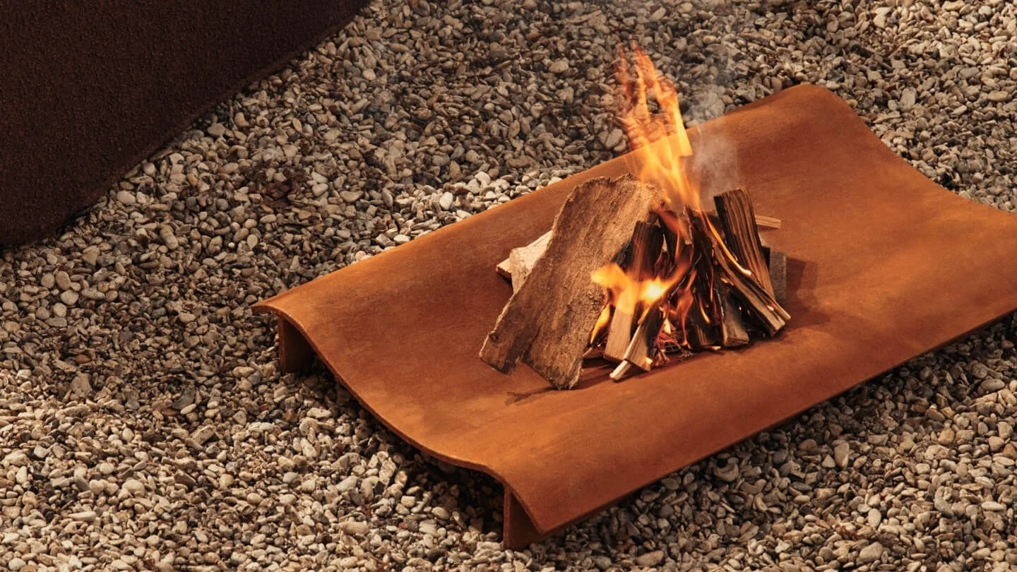Pequeña hoguera con leña ardiendo en una bandeja metálica rectangular oxidada sobre grava.