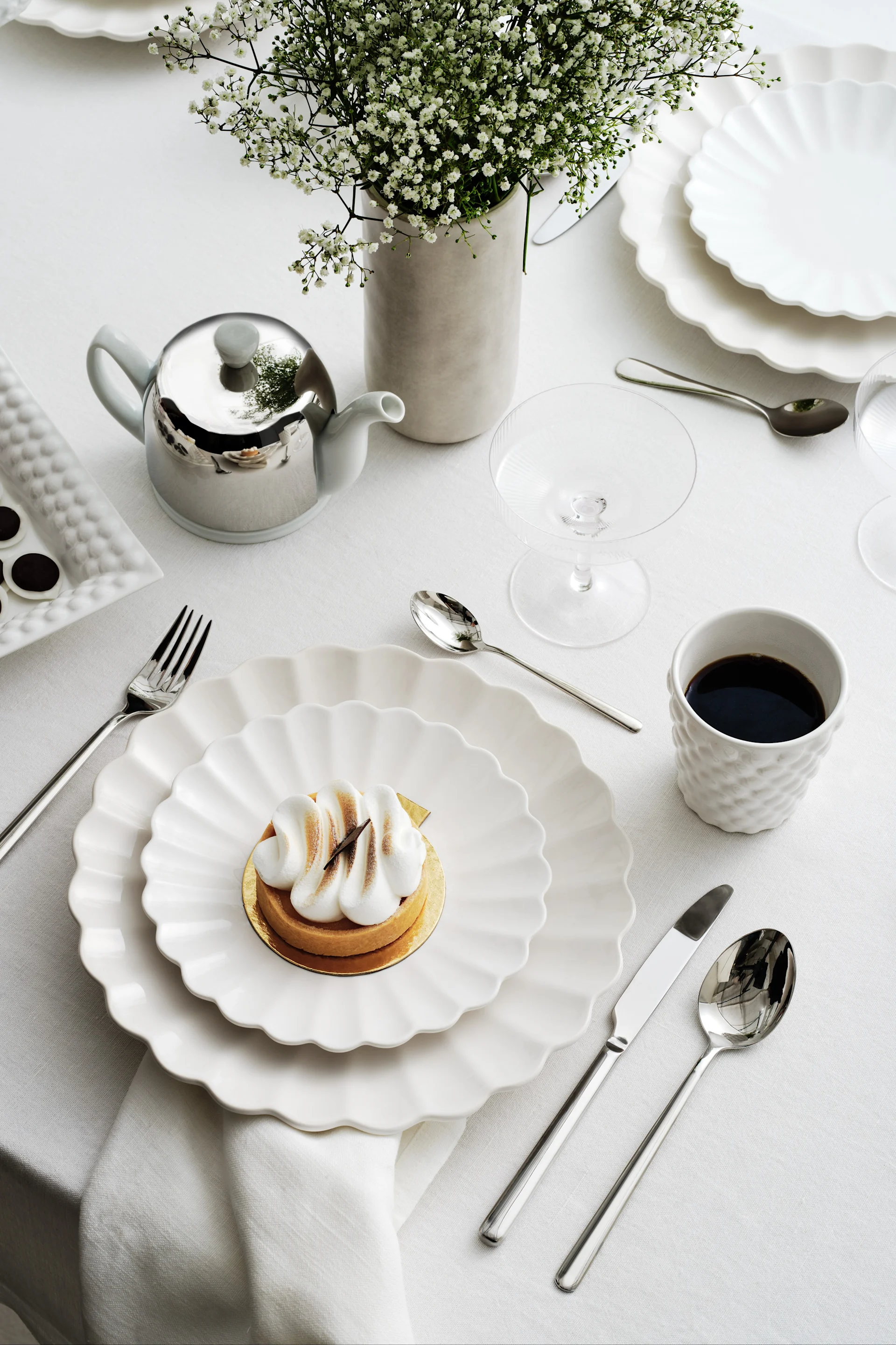 Mesa elegante con platos blancos festoneados, una tarta de merengue, café, una tetera plateada y flores de paniculata.