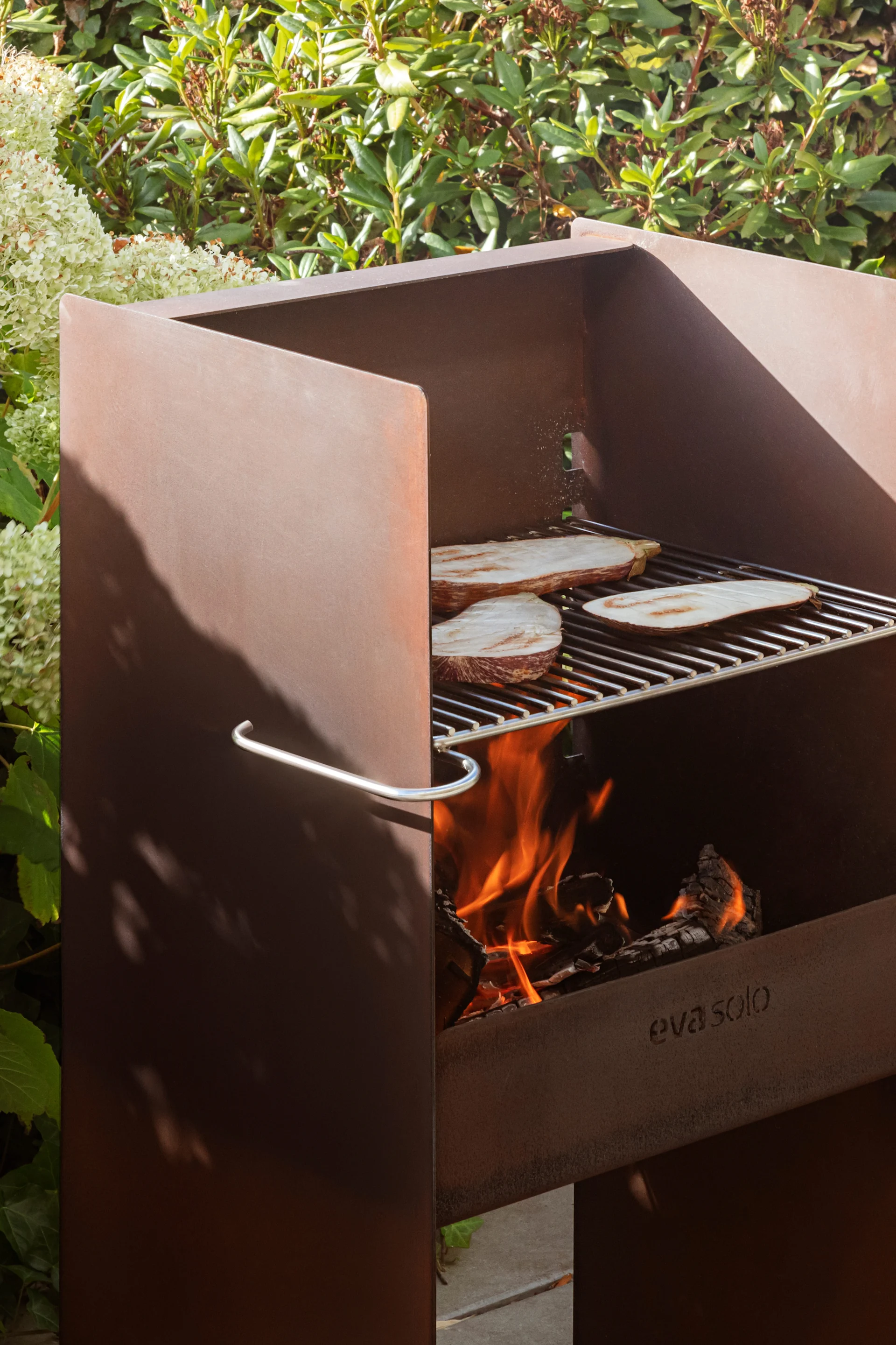 Parrilla de exterior de acero corten con rodajas de berenjena cocinándose sobre una rejilla y fuego abierto, rodeada de plantas verdes.