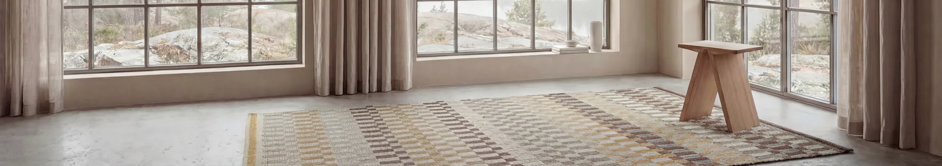 Una habitación con grandes ventanales que revelan un paisaje rocoso. Una alfombra estampada y un taburete de madera clara sobre un suelo gris, con decoración en el alféizar.