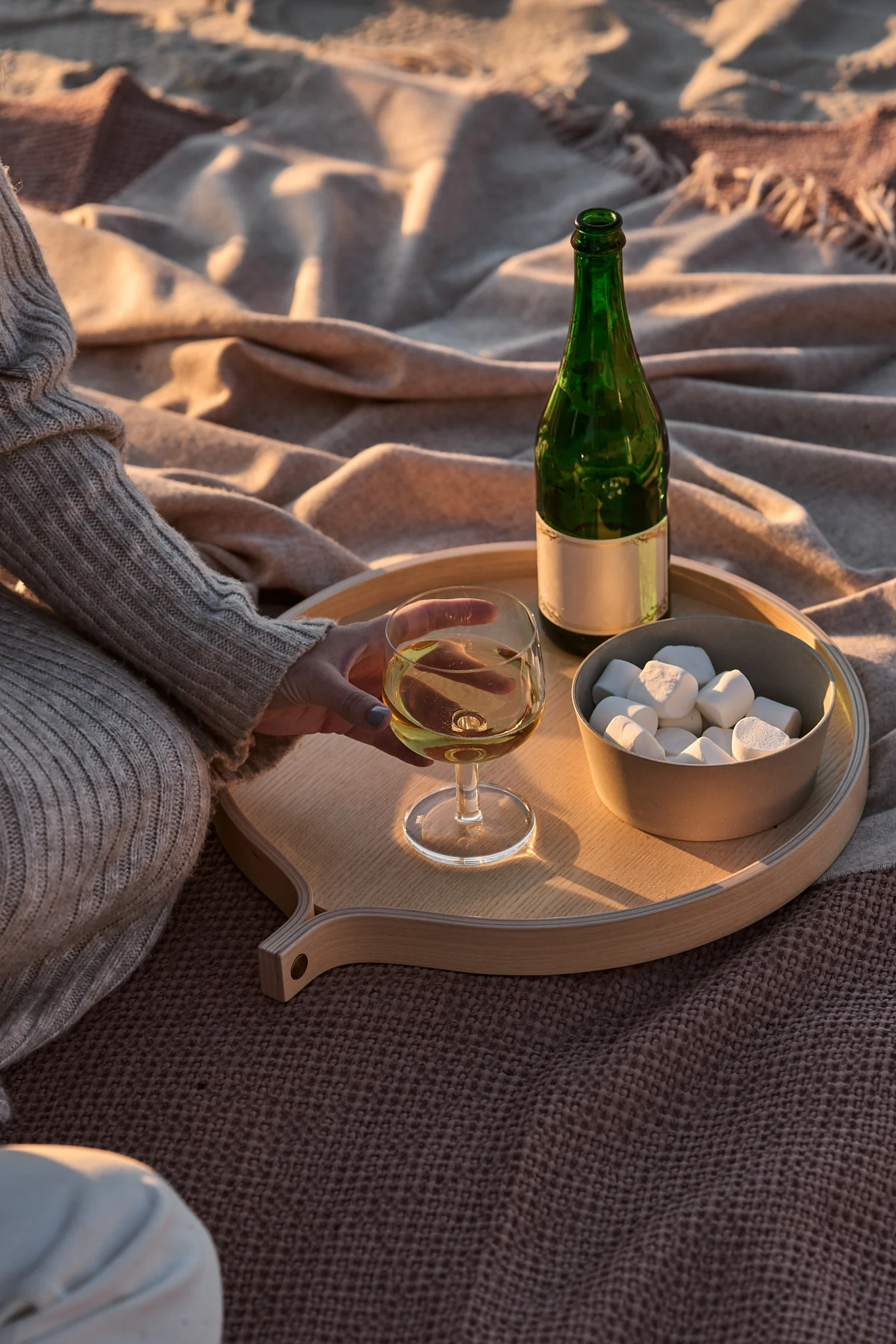Persona sosteniendo vino blanco en bandeja de madera con botella verde y malvaviscos sobre mantas al atardecer.