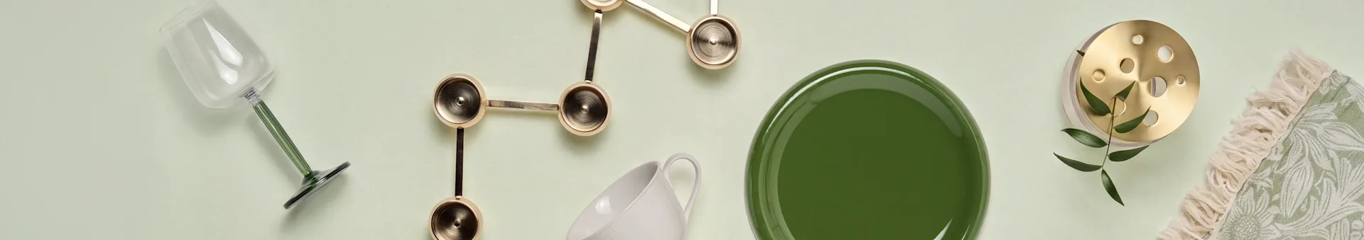 Vista superior: copa con tallo verde, adorno dorado escultórico, taza blanca, plato verde, objeto de latón con hojas y textil verde con flecos.