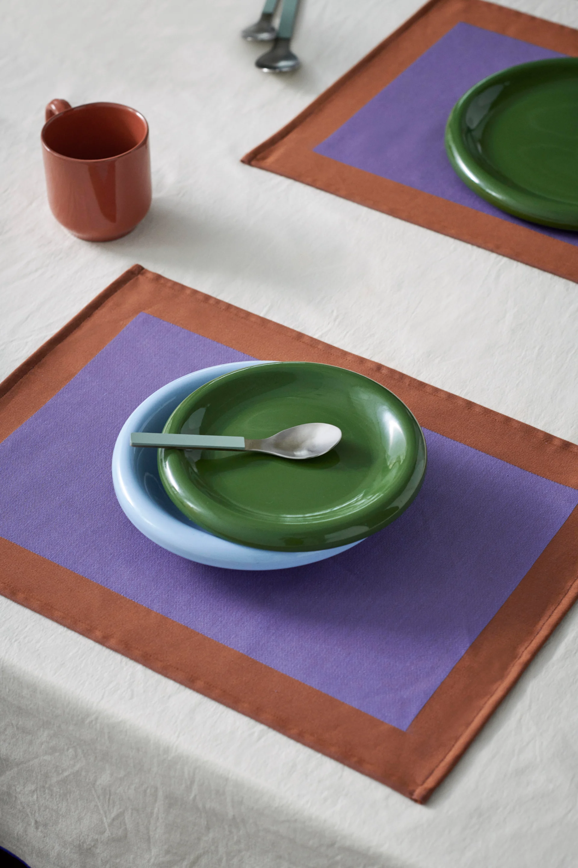 Servicio de mesa con cuenco azul, plato verde y cuchara en mantel individual morado y marrón. Una taza marrón en el mantel blanco.