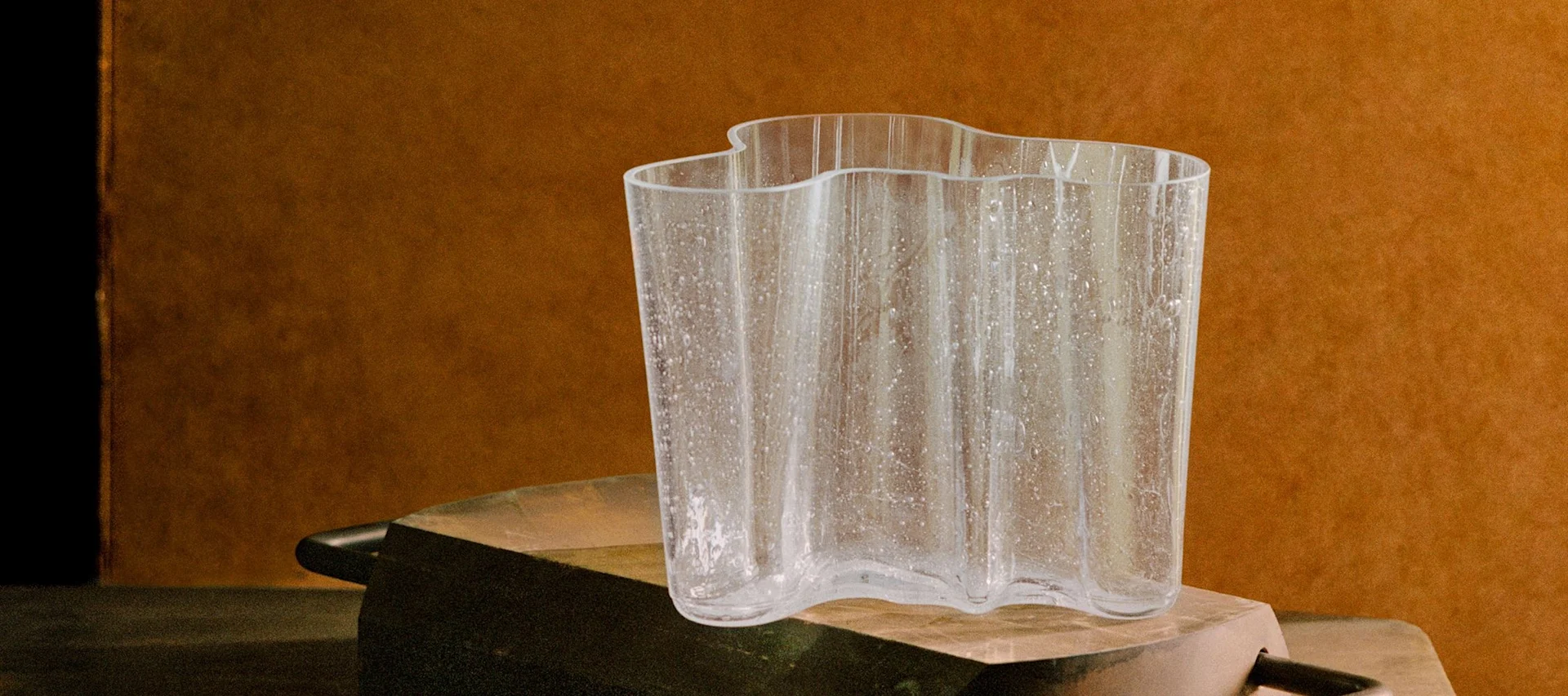 Un jarrón de vidrio transparente y ondulado con motas texturizadas sobre una superficie rústica oscura y un fondo marrón cálido.
