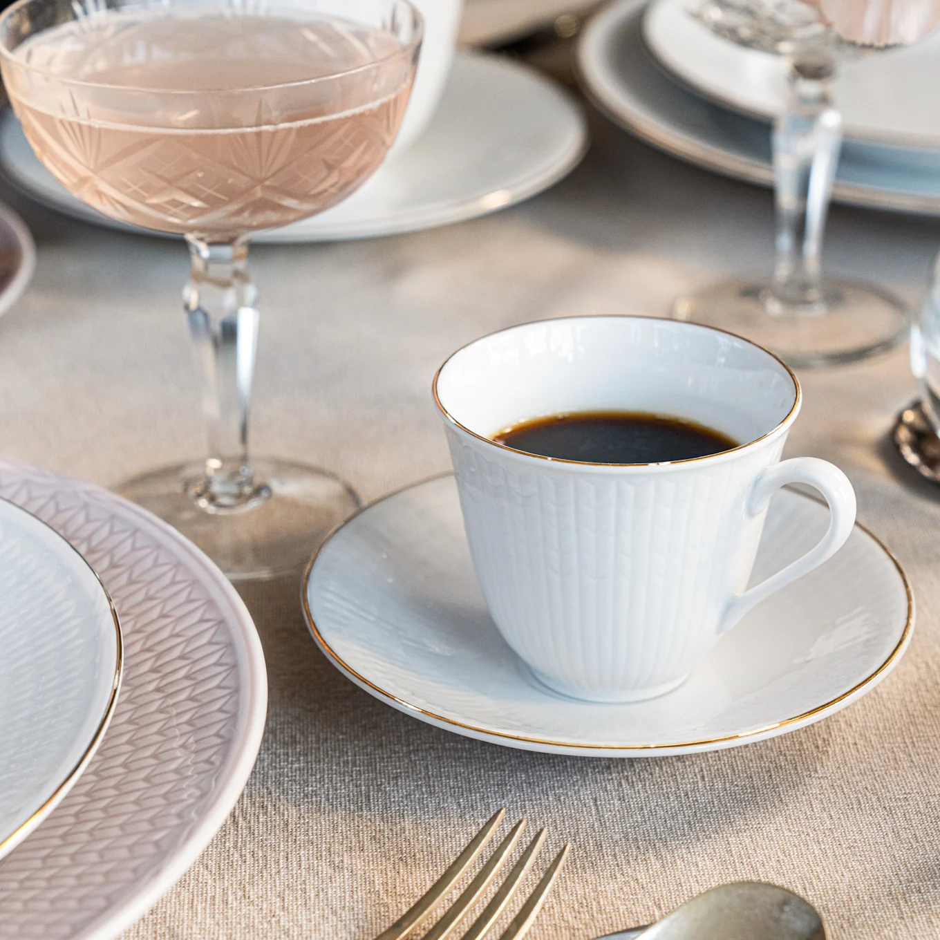 Taza de café blanca con borde dorado sobre un platito, junto a un plato rosa y una copa rosada sobre una mesa de lino.