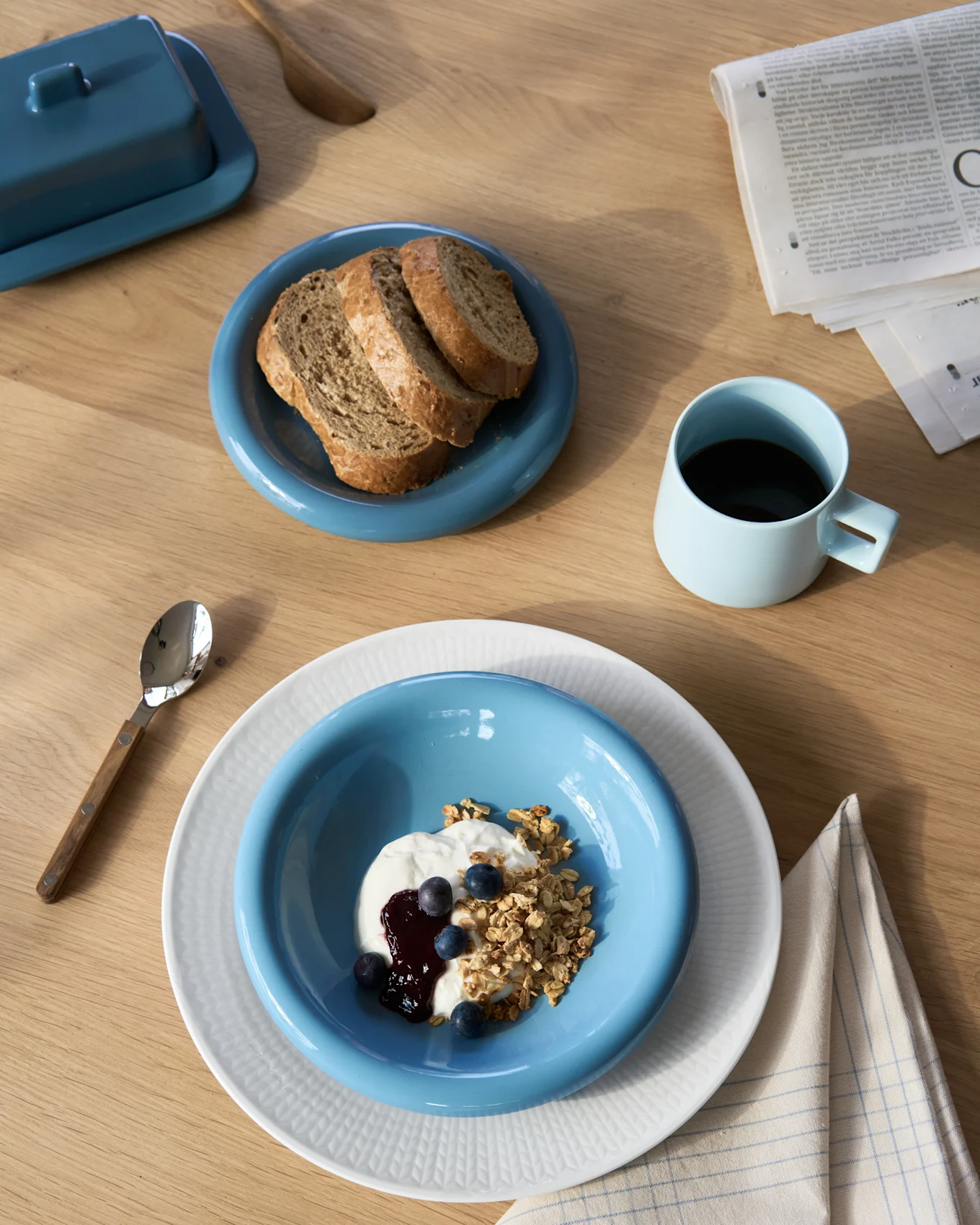 Vista aérea de un desayuno con yogur y granola, pan integral, café y una mantequillera sobre una mesa de madera.