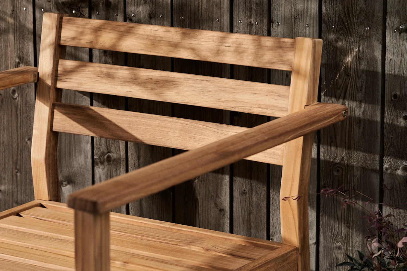 Silla de madera con lamas y reposabrazos contra una valla de madera oscura, cubierta de sombras de hojas.
