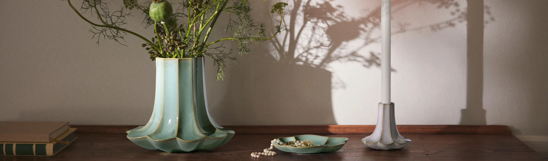 Un jarrón estriado de color verde azulado con follaje, un collar de perlas en un plato a juego, una vela blanca en un candelabro estriado y libros sobre una superficie de madera.