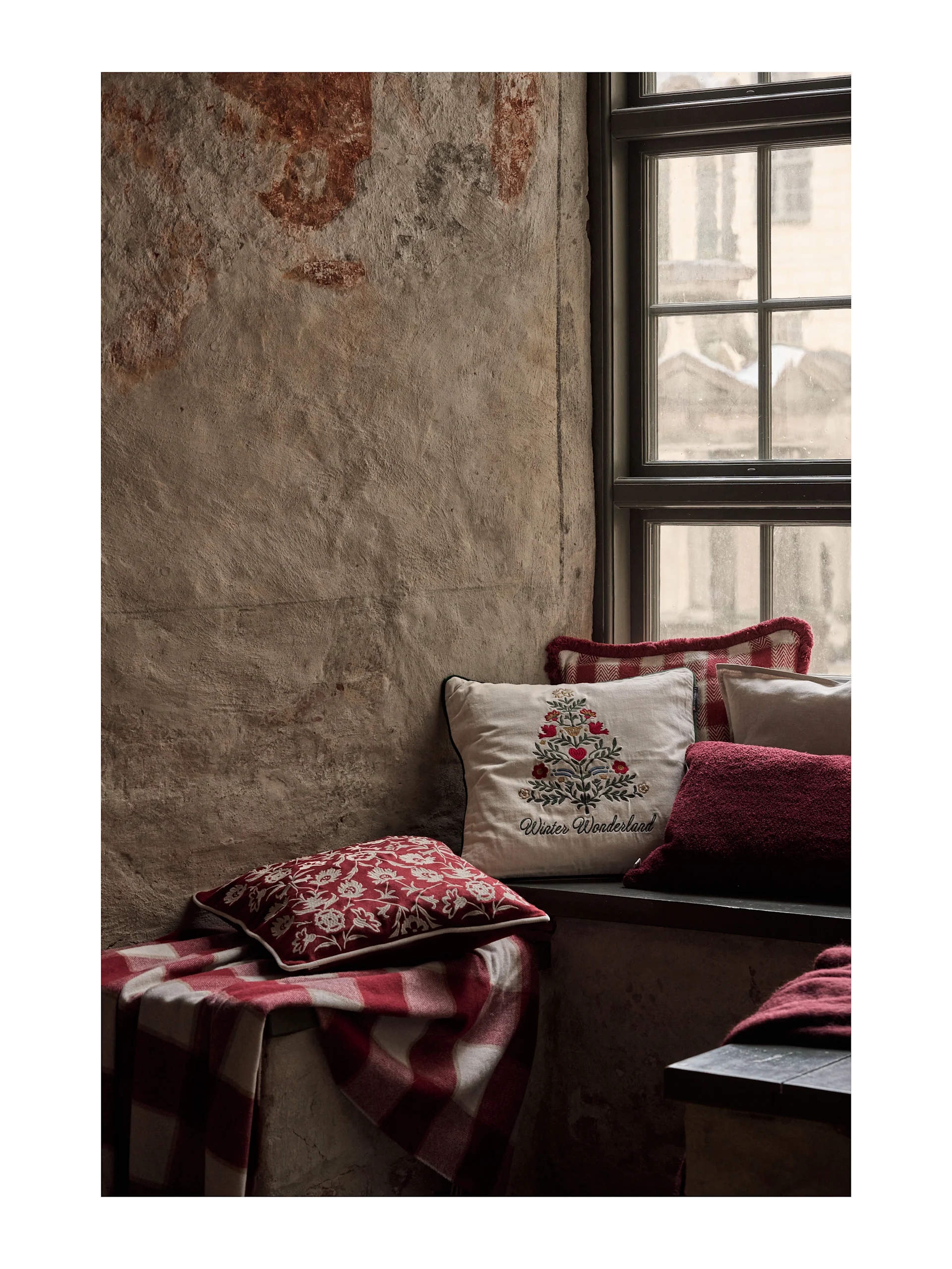 Acogedor asiento junto a la ventana con cojines festivos rojos y blancos y una manta de cuadros. Un cojín crema lleva el bordado 'Winter Wonderland'.