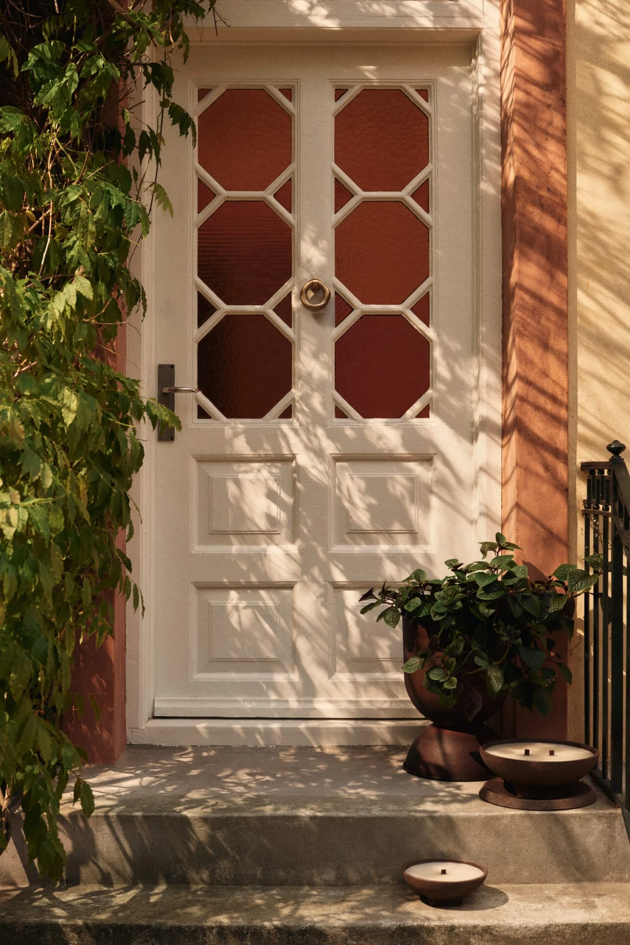 Puerta principal blanca con paneles de vidrio octogonales, enredaderas verdes a la izquierda y una maceta con dos velas en los escalones.
