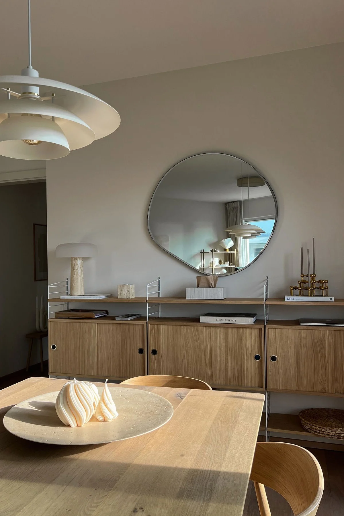 Comedor moderno con mesa de madera, dos velas en espiral, estante, espejo irregular y lámpara blanca escalonada.